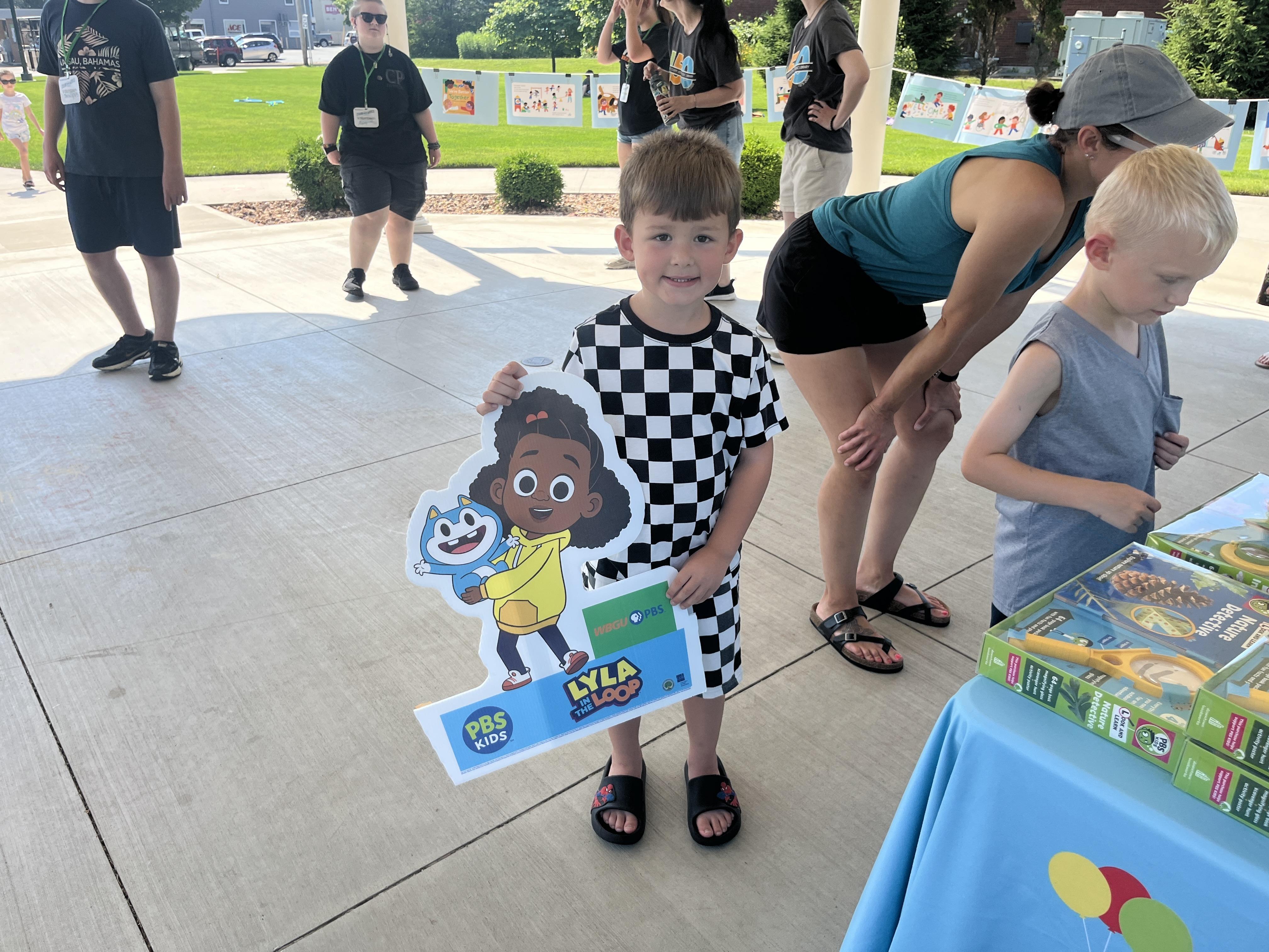 A young boy in a black and white checkered t-shirt smiles at the camera, holding a cutout sign featuring characters from "Lyla in the Loop" and the PBS Kids logo. He is standing on a paved outdoor area. In the background, several adults and children are visible, and a table covered with a light blue tablecloth featuring WBGU-PBS branding can be seen to the right. The setting appears to be an outdoor event on a sunny day.
