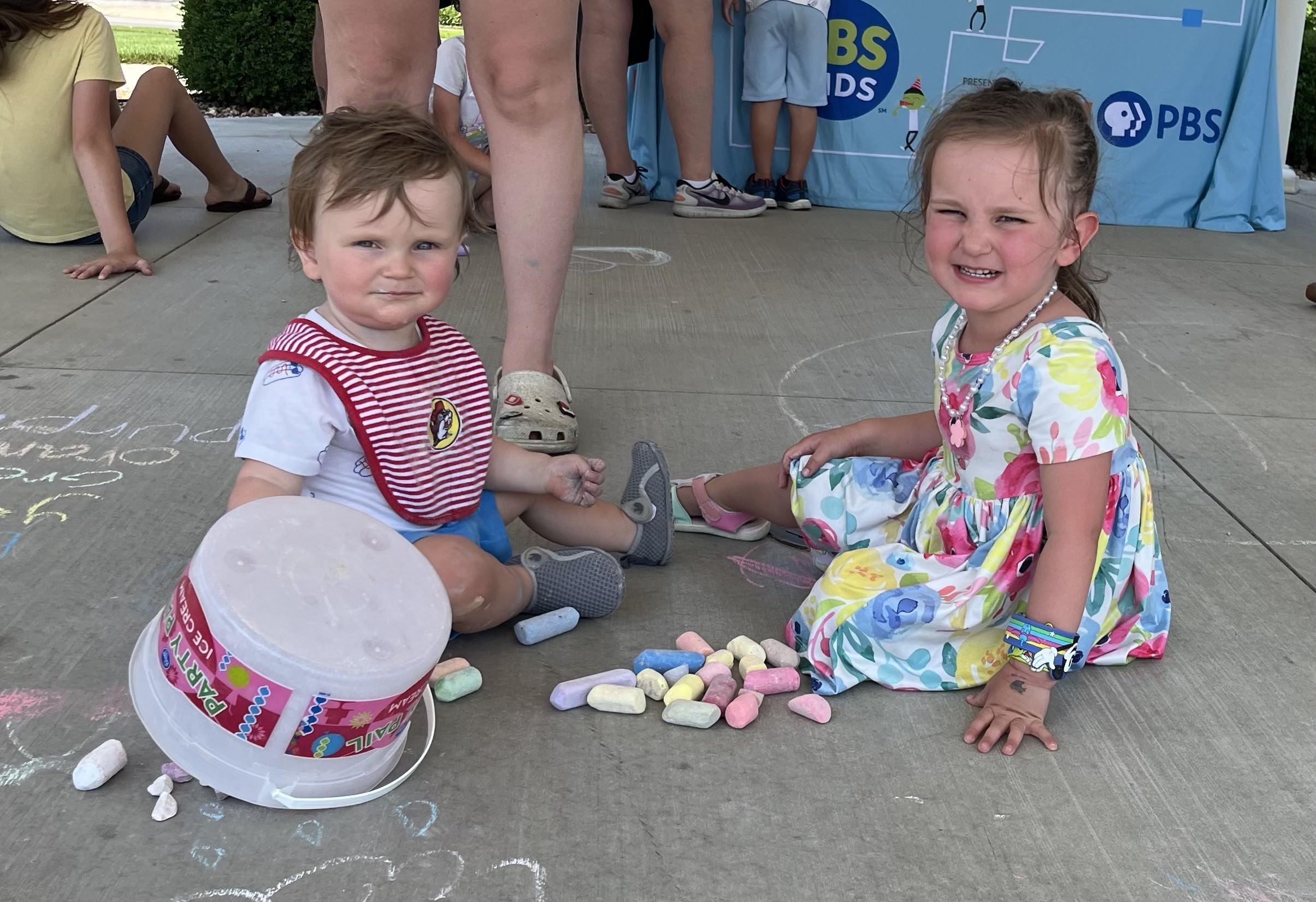  Two young children are seated on a paved surface, surrounded by colorful sidewalk chalk. The child on the left, a toddler wearing a red and white striped bib over a white shirt and blue shorts, looks towards the viewer.