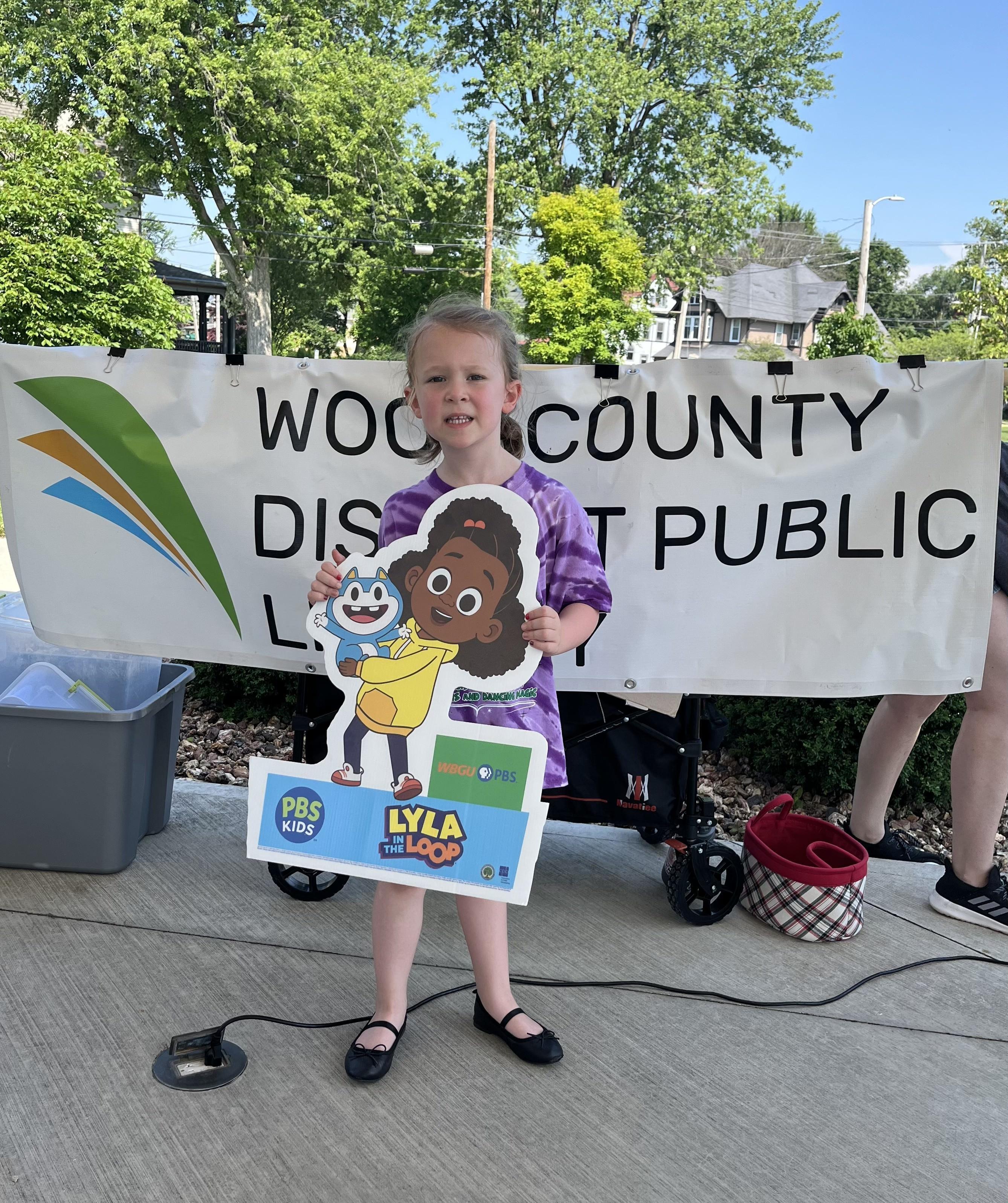 A young girl with a slight smile and light brown pigtails, wearing a purple tie-dye shirt and black flats, holds a "Lyla in the Loop" and PBS Kids sign. She stands in front of a large white banner that says "WOOD COUNTY DISTRICT PUBLIC LIBRARY". The setting is outdoors in Bowling Green, Ohio, with trees and a house visible in the background.