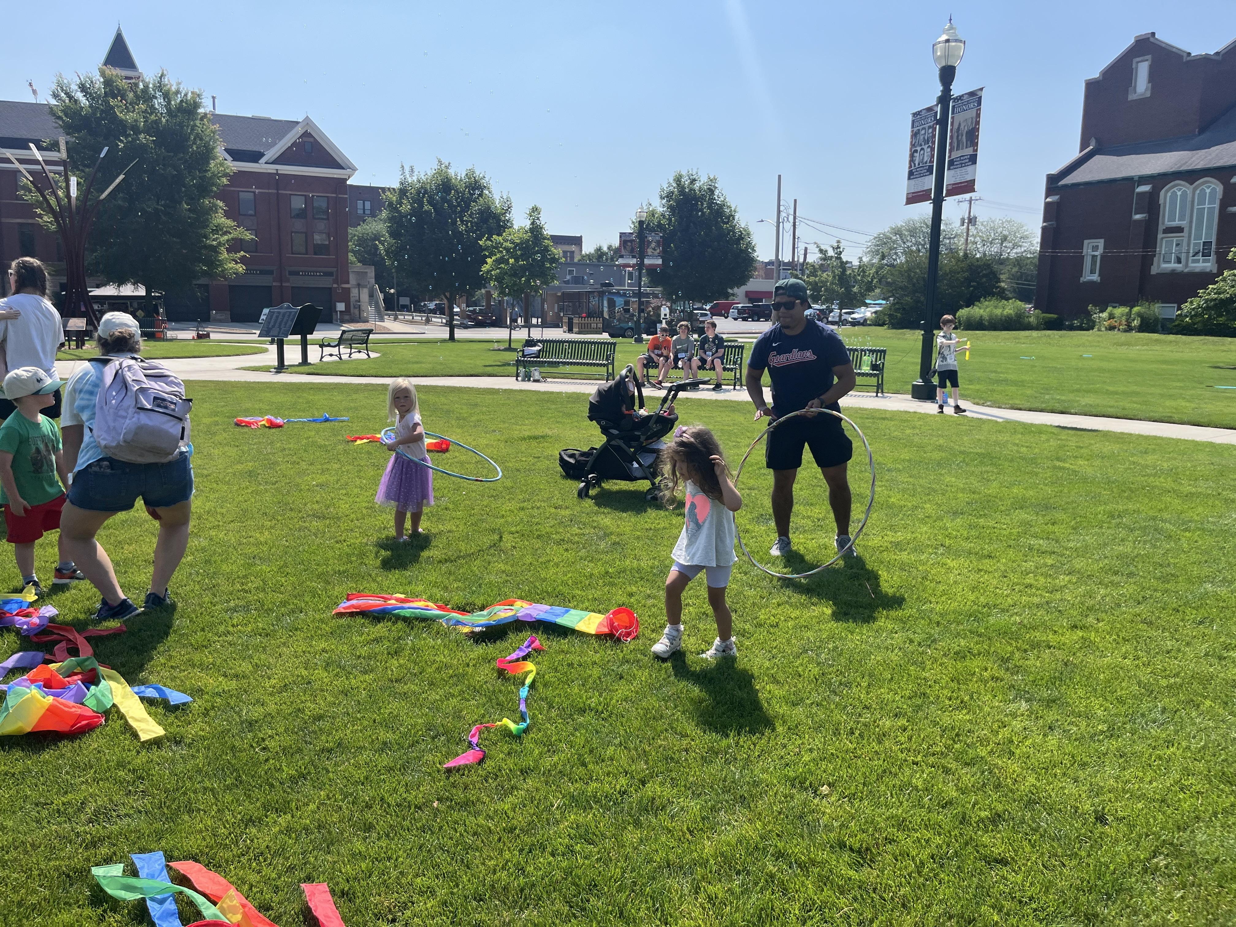 A vibrant outdoor scene on a sunny day in Bowling Green, Ohio, shows several children and adults enjoying a grassy park. In the foreground, two young girls are playing with hula hoops and colorful fabric strips laid on the bright green lawn. To the left, an adult and another child are near a pile of colorful items. In the midground, other people are visible, including a man in shorts and a dark shirt standing with a hula hoop, and others sitting on benches. Beyond the park, historic-looking buildings and trees are visible under a clear blue sky.