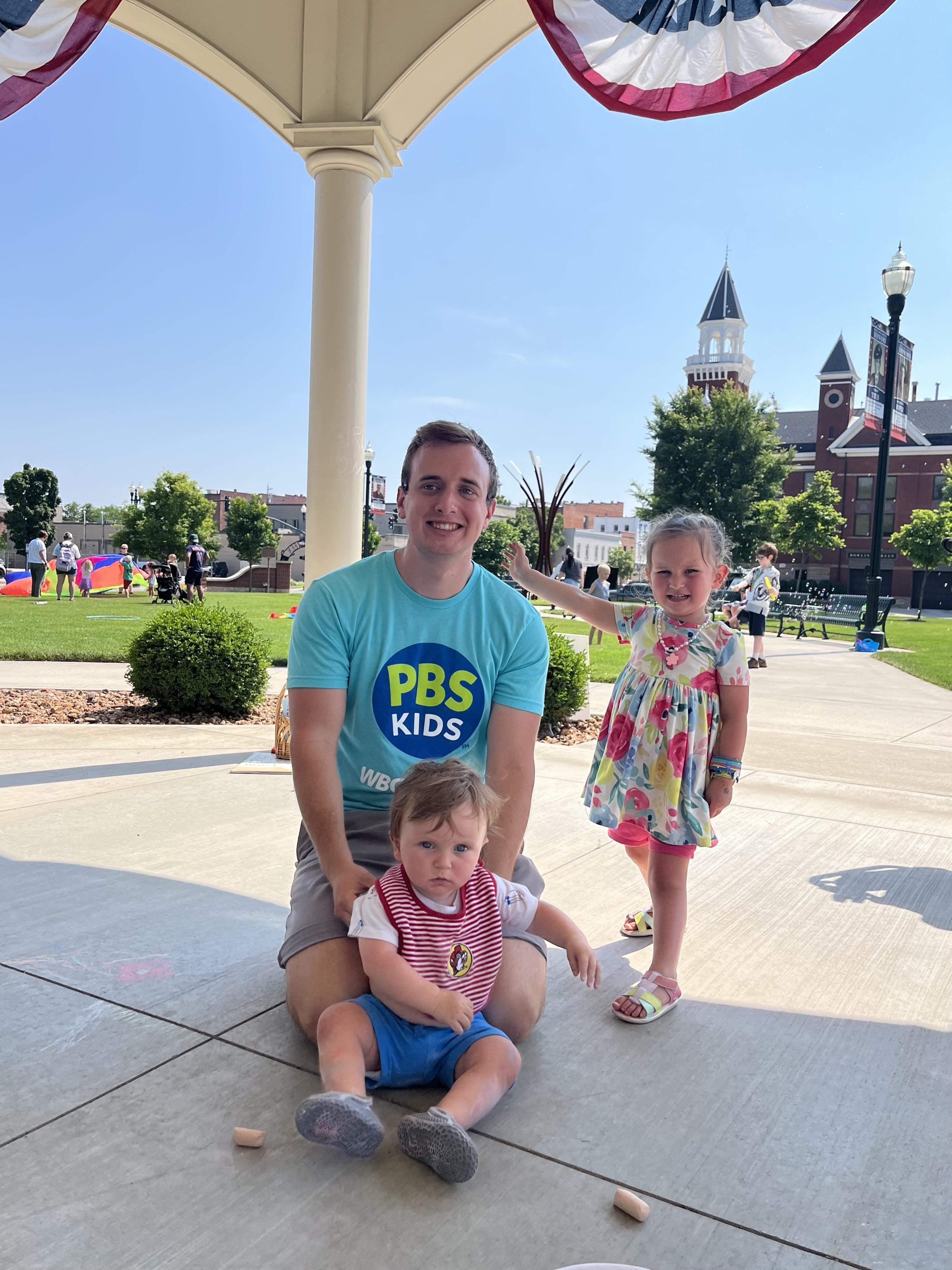 A smiling man wearing a teal PBS Kids t-shirt kneels on a paved area, with a baby in a red and white striped shirt and blue shorts sitting in front of him. To the right, a young girl in a floral dress stands beside them, smiling. They are under a canopy decorated with red, white, and blue bunting. In the background, a sunny park with people and a distinctive building with a steeple are visible in downtown Bowling Green, Ohio.