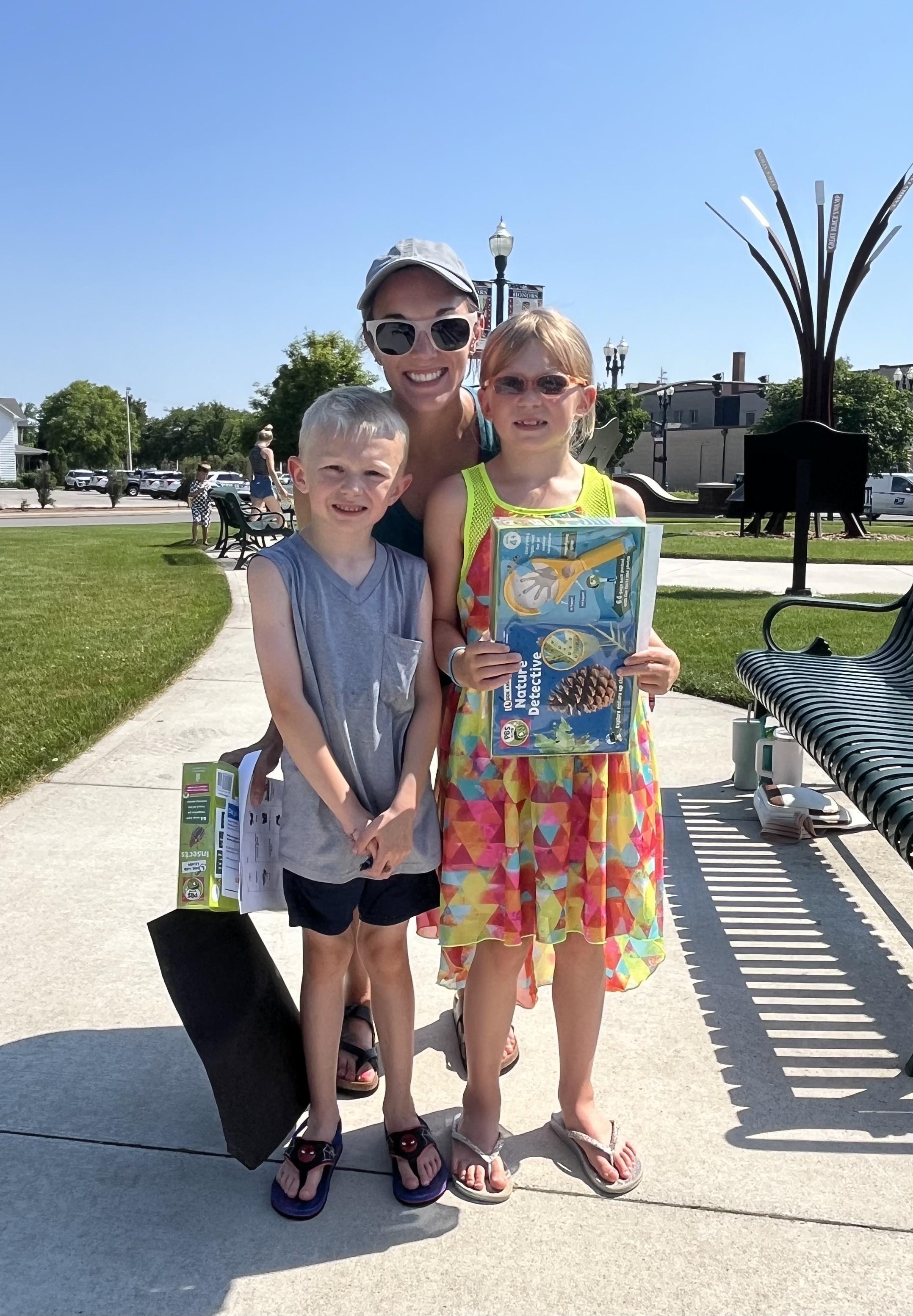 A smiling adult woman wearing a hat and sunglasses stands between two children on a paved path outdoors. To her left, a boy in a grey t-shirt and dark shorts smiles at the camera. To her right, a girl with sunglasses on her head, wearing a colorful patterned dress, holds up a "Nature Detective" book. They appear to be at an event in Bowling Green, Ohio, with a park setting, benches, and buildings visible in the background under a clear sky.
