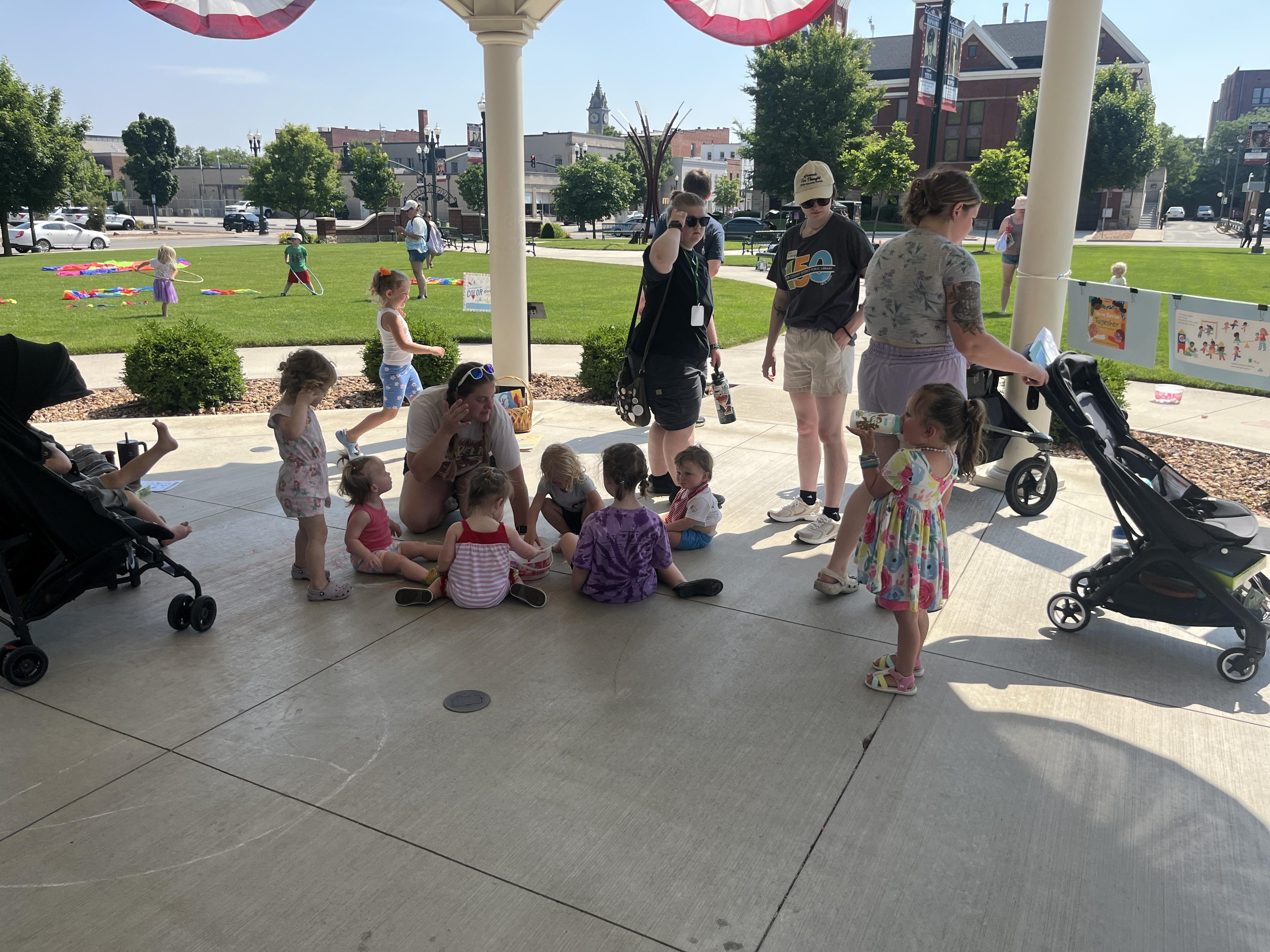 A lively outdoor scene shows several young children and adults gathered on a paved area under a canopy. In the foreground, a group of toddlers are sitting or kneeling around an adult, while another toddler stands near a black stroller. In the background, on a sunny green lawn, more children and adults are spread out, with some engaging in activities like hula-hooping. Buildings and trees are visible in the distance under a clear sky.