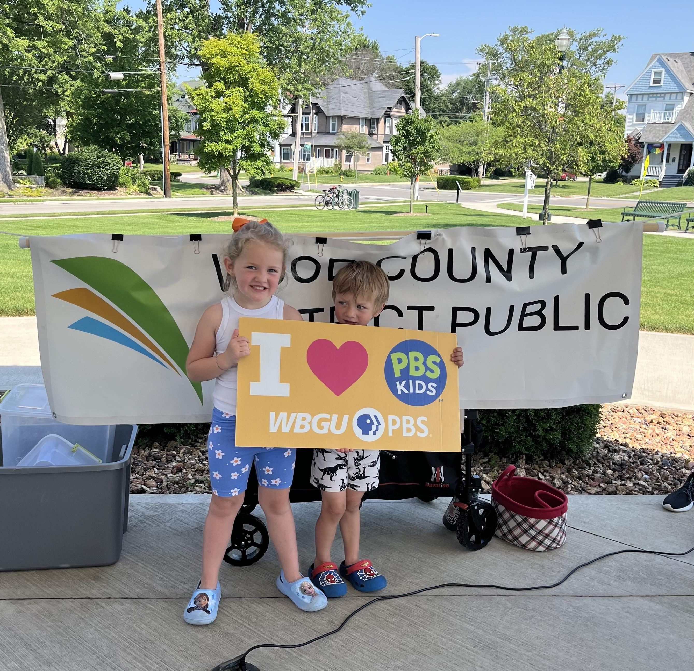 Two young children are holding a yellow sign that reads "I [heart symbol] PBS KIDS WBGU PBS" in front of a large white banner for the "WOOD COUNTY DISTRICT PUBLIC LIBRARY". The girl on the left has blonde pigtails and is wearing a white tank top and blue patterned shorts with light blue clogs. The boy on the right is wearing patterned shorts and blue clogs, peeking from behind the sign. They are standing outdoors on a paved surface, with trees and residential buildings visible in the background under a clear sky in Bowling Green, Ohio.