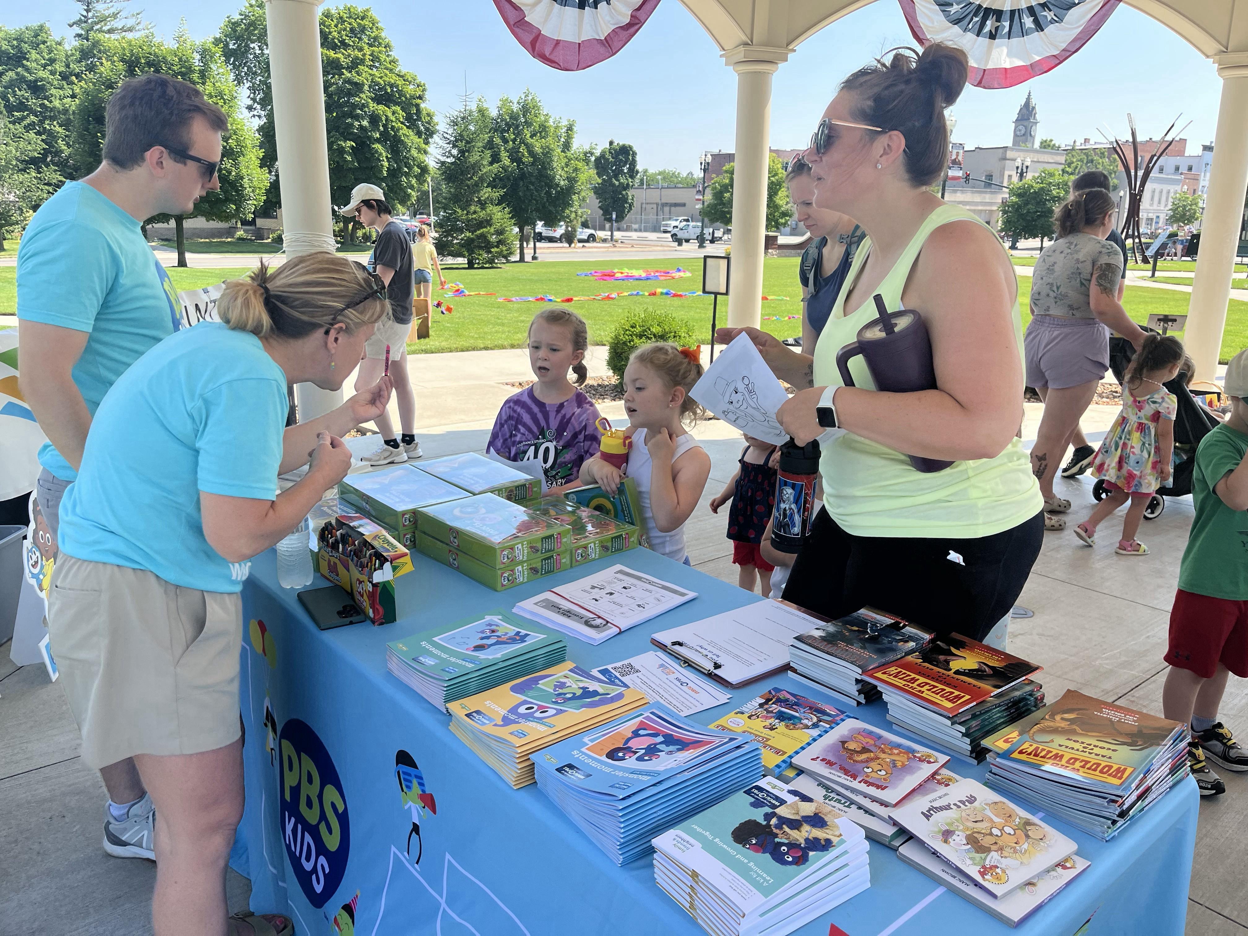 Under a white gazebo with red, white, and blue bunting, two adults in teal PBS Kids shirts are interacting with children at a table. The table is covered with a light blue tablecloth branded "PBS Kids" and holds stacks of books, green boxes, and papers. Two young girls are looking at the table's contents. In the background, other people are visible on a sunny green lawn, and buildings of downtown Bowling Green, Ohio, can be seen in the distance.
