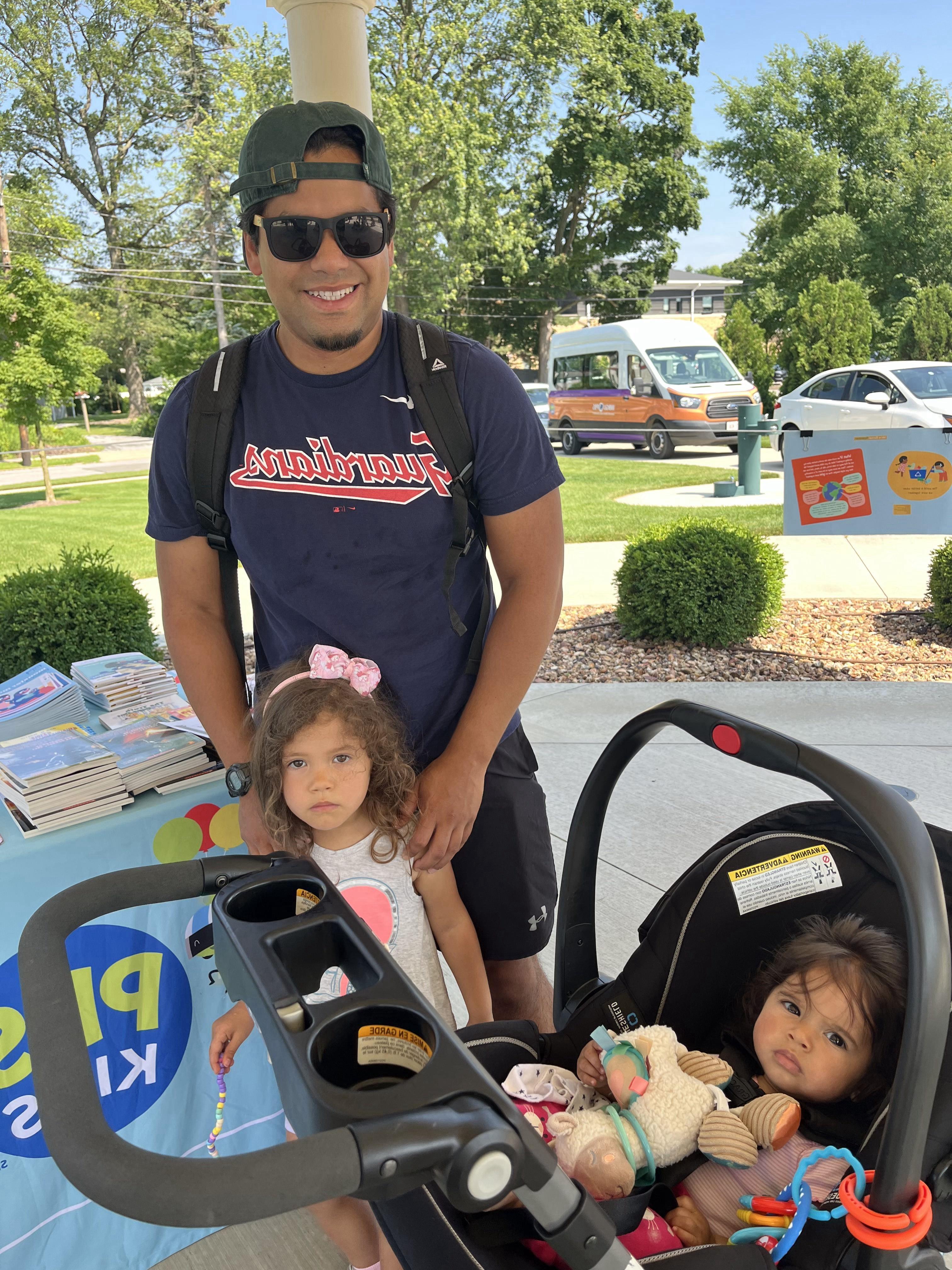 A smiling man wearing a dark blue "Guardians" t-shirt, a baseball cap, and sunglasses stands next to a baby in a car seat and a young girl with curly hair and a pink bow. The baby is looking towards the left, while the girl is looking towards the camera. To the right, part of a light blue table with "PBS Kids" branding and stacks of books is visible. The scene is outdoors on a sunny day in Bowling Green, Ohio, with trees and a white van in the background.