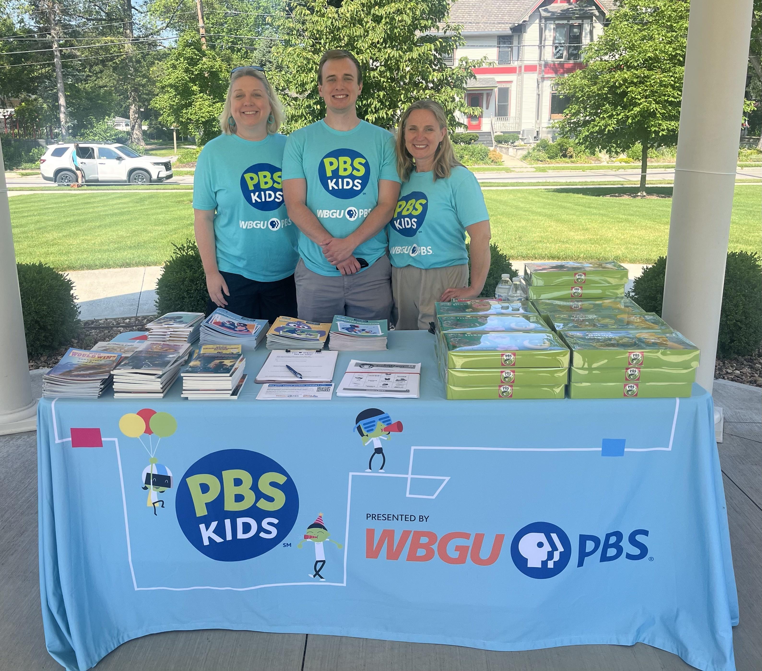 Three adults, two women and one man, all smiling and wearing teal PBS Kids t-shirts, stand behind a light blue table covered with a "PBS Kids Presented by WBGU PBS" tablecloth. The table is laden with books, papers, and stacks of green boxes. The setting is outdoors under a canopy or portico, with a green lawn and residential buildings visible in the background in Bowling Green, Ohio, on a sunny day.