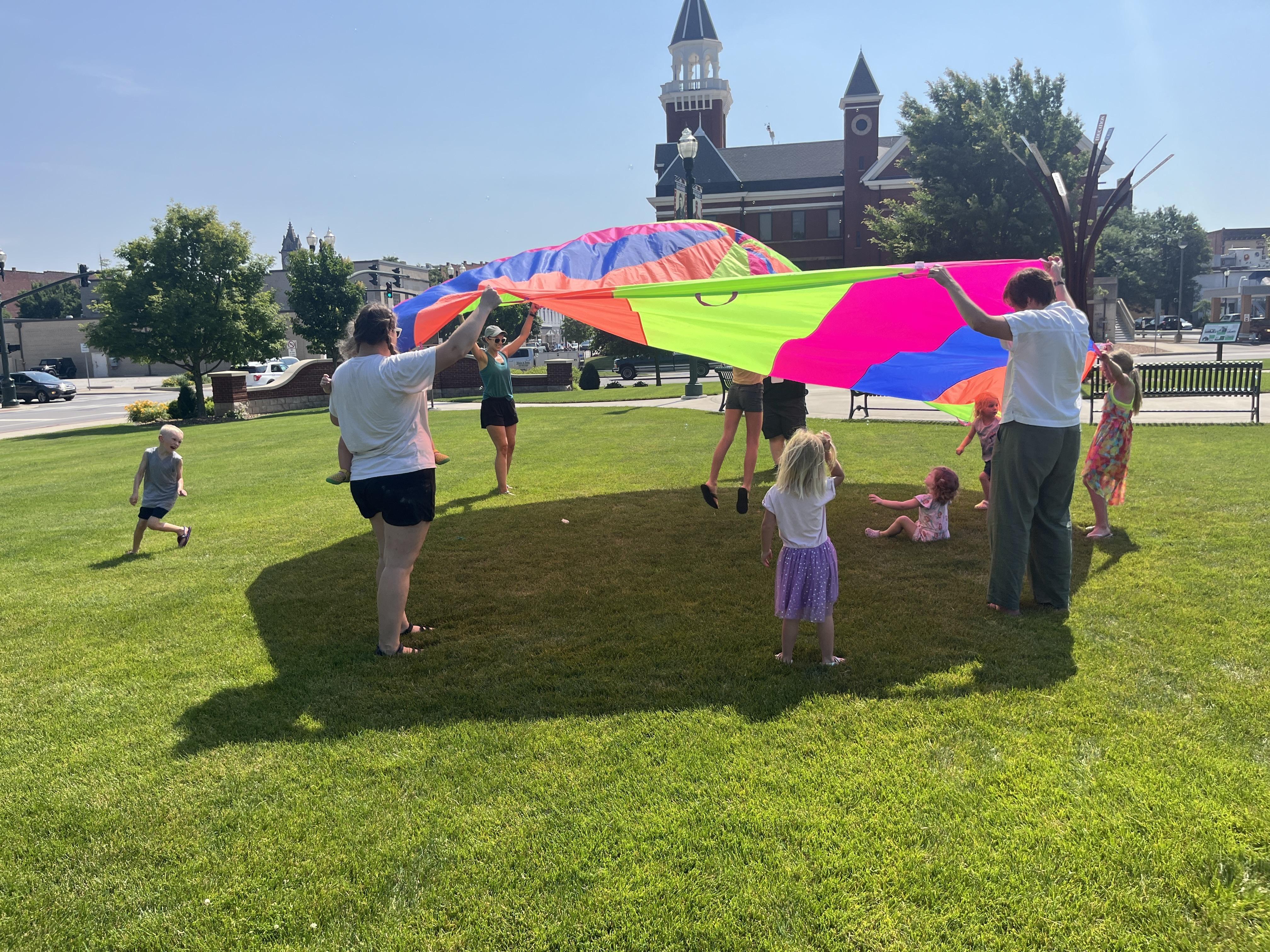 On a sunny day in Bowling Green, Ohio, a group of children and adults are playing with a large, colorful parachute on a bright green lawn. The parachute, featuring panels of pink, green, orange, and blue, is held aloft by several adults and children. Other children are visible under or near the parachute, some sitting on the grass. In the background, a distinctive building with a steeple and other structures of downtown Bowling Green are visible under a clear blue sky, along with trees and benches.