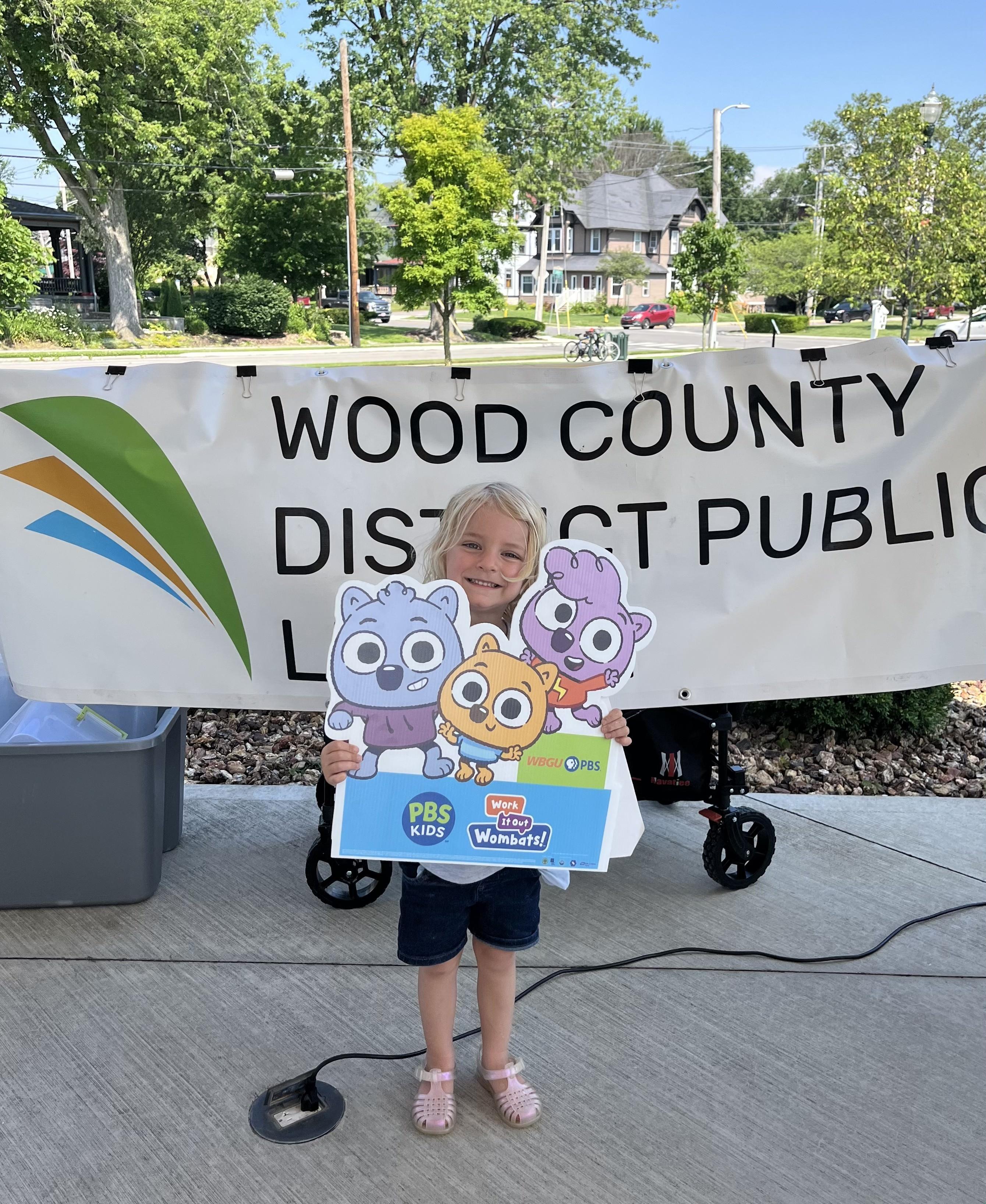 A young girl with blonde hair, wearing a white t-shirt and blue shorts, smiles brightly as she holds up a colorful sign featuring "Work It Out Wombats!" characters and PBS Kids logos. She is standing outdoors in front of a large white banner that partially reads "WOOD COUNTY DISTRICT PUBLIC LIBRARY." In the background, trees and residential buildings are visible under a clear sky.