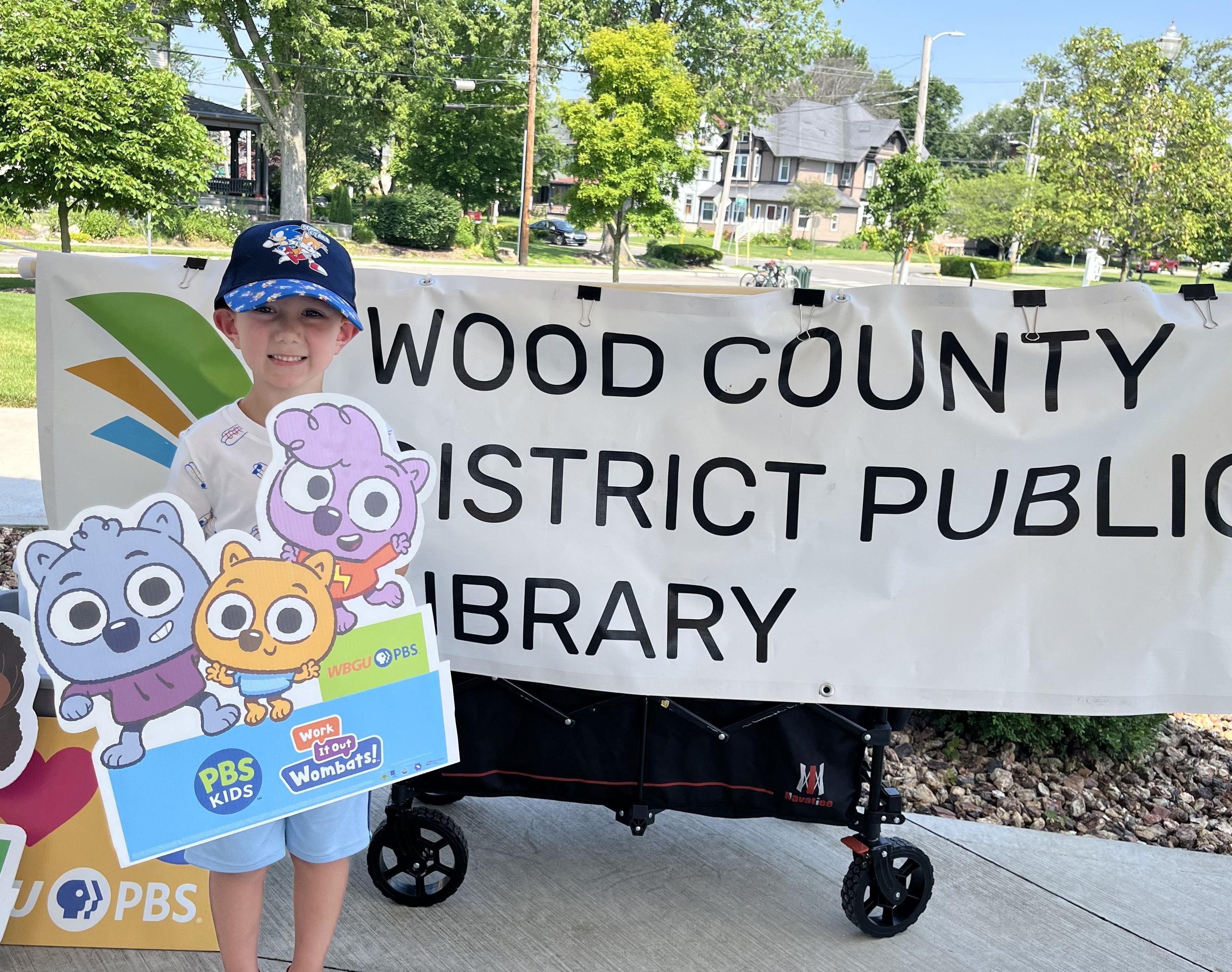 A young boy, smiling and wearing a blue and white baseball cap, stands in front of a large white banner that reads "WOOD COUNTY DISTRICT PUBLIC LIBRARY." He is holding a colorful sign featuring cartoon characters from "Work It Out Wombats!" and PBS Kids logos. To his right, a black wagon is visible. The scene is outdoors with trees, houses, and a sunny sky in the background.