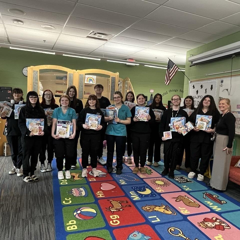 The PENTA students are pictured in their preschool classroom with their instructor, Mrs. Dova Zak, holding some of the items they received including Wild Kratts Board Games and children's books. .