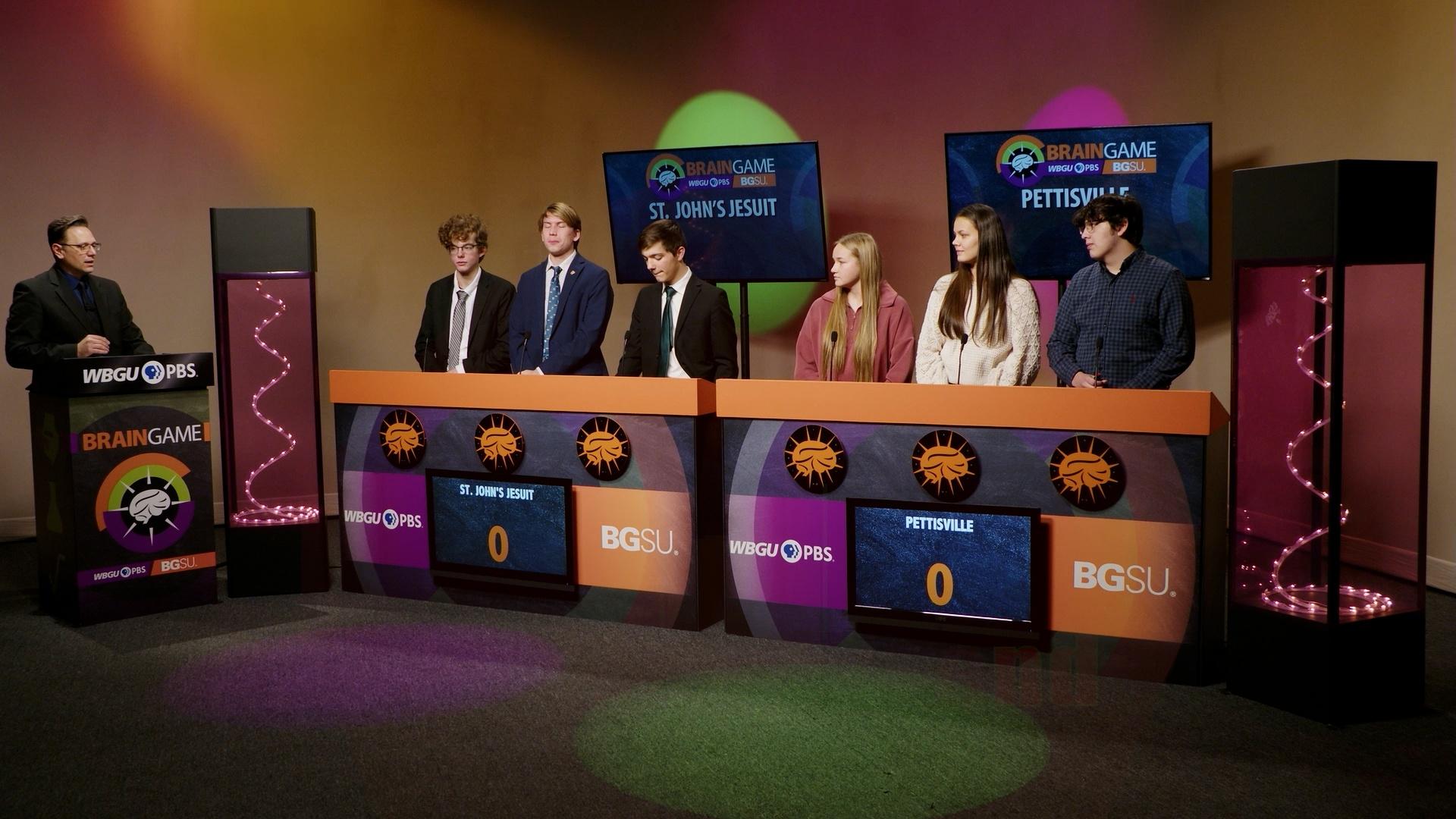 An image from the BGSU Brain Game set. Host Marc Hathaway is behind the podium on the left side and in the center there are two larger podiums- one for St. John’s Jesuit and one for Pettisville. There are three students behind each of the larger podiums. Green and purple lights adorn the set.