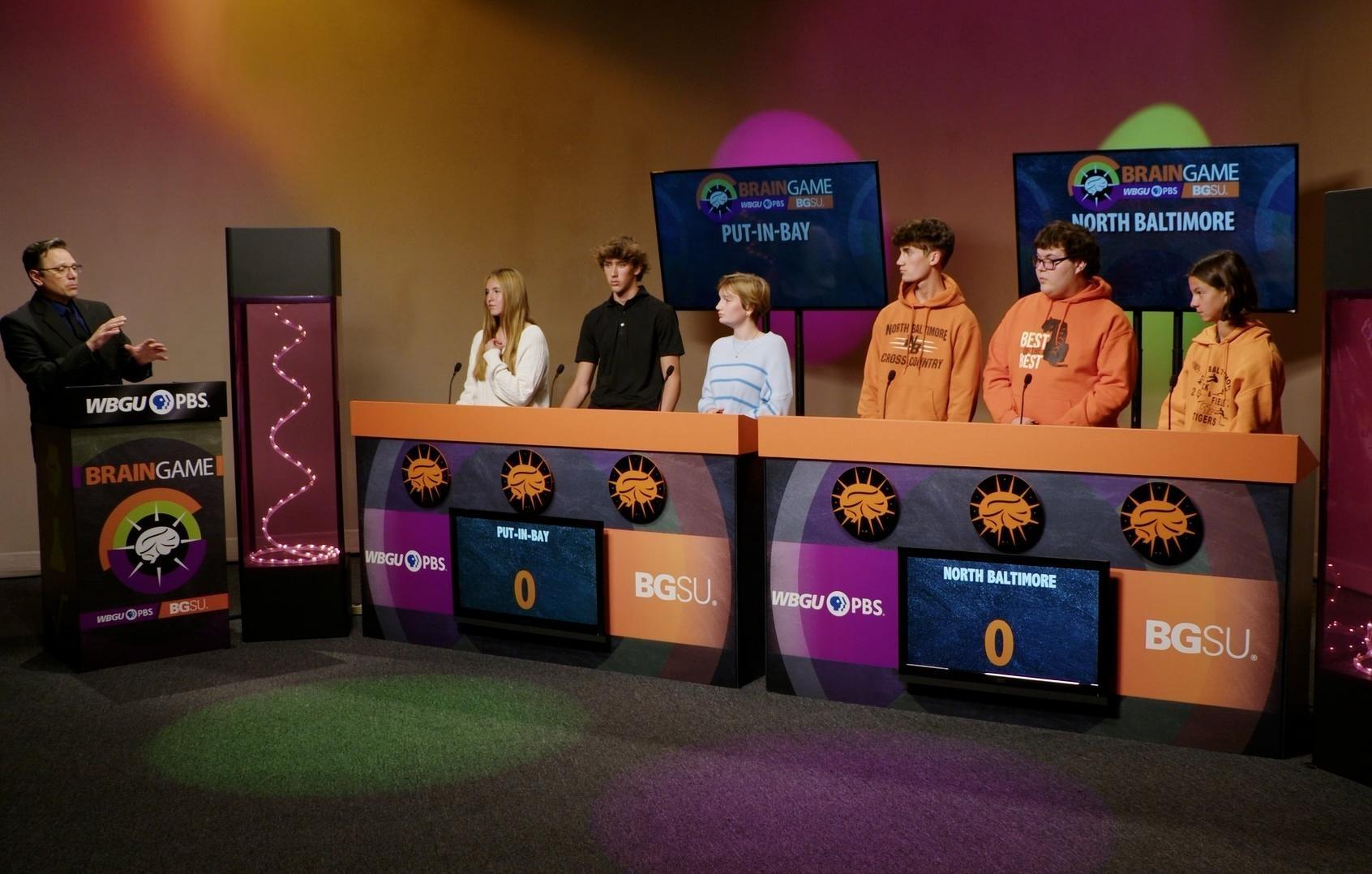 Two quiz bowl teams on the set of BGSU Brain Game. Host Marc Hathaway is behind a podium on the far left. 3 players are behind a podium that says "Put-in-Bay" and next to them are 3 players behind a podium that says "North Baltimore"