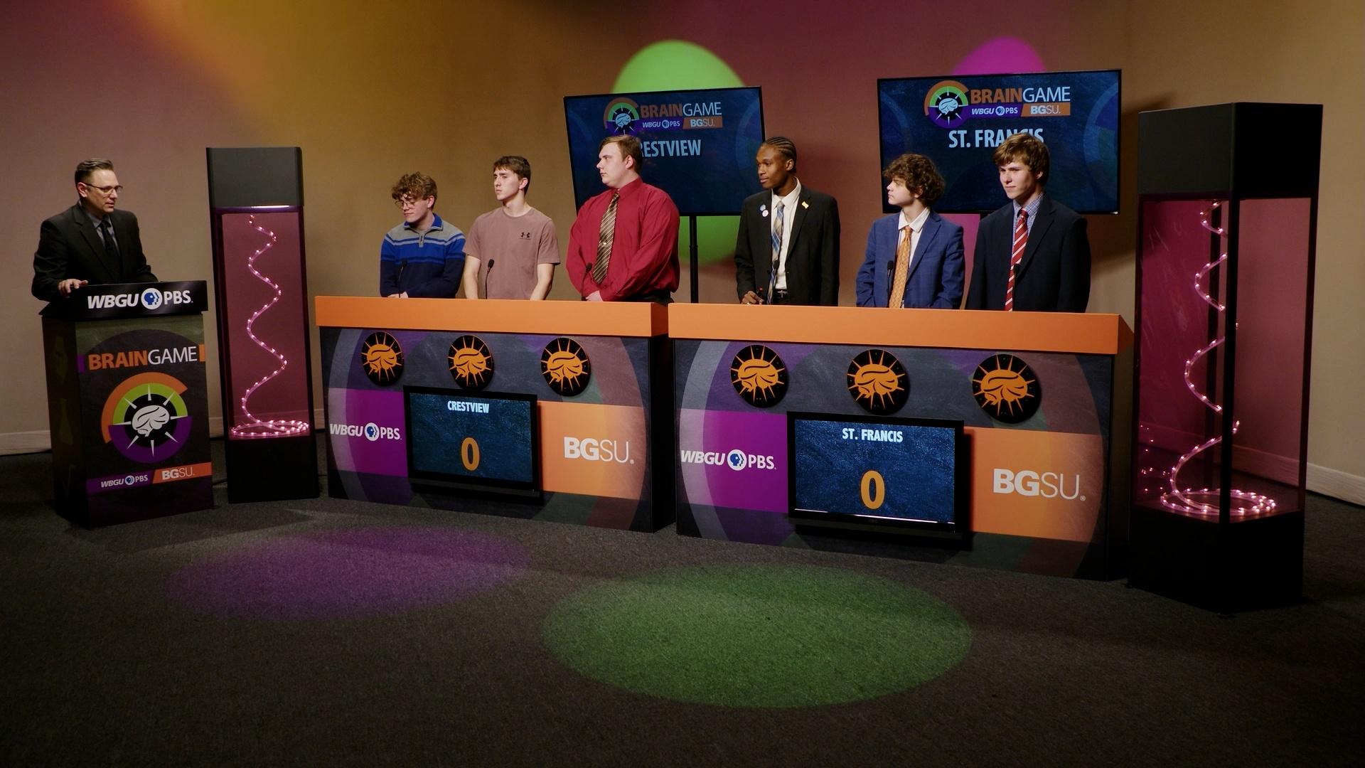 An image from the BGSU Brain Game set. Host Marc Hathaway is behind the podium on the left side and in the center there are two larger podiums- one for Crestview and one for St. Francis. There are three students behind each of the larger podiums. Green and purple lights adorn the set.