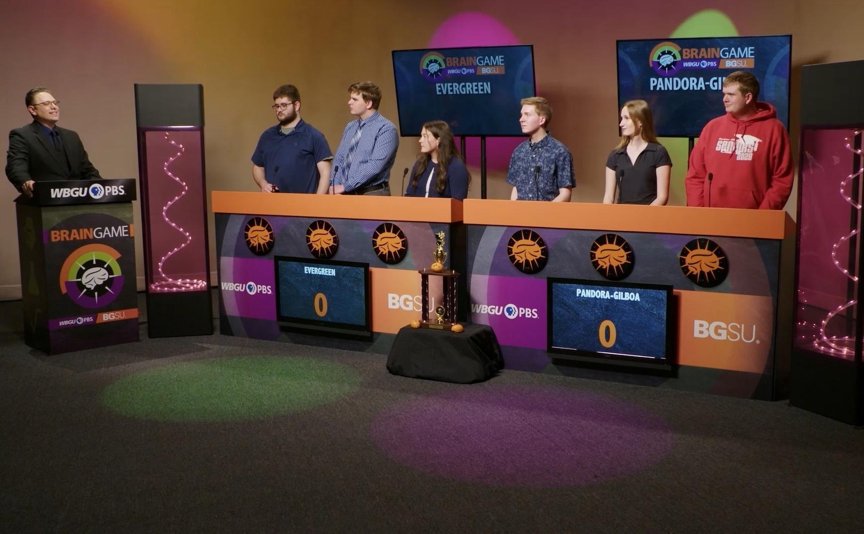 An image from the BGSU Brain Game set. Host Marc Hathaway is behind the podium on the left side and in the center there are two larger podiums- one for Evergreen and one for Pandora-Gilboa. There are three students behind each of the larger podiums. Green and purple lights adorn the set.