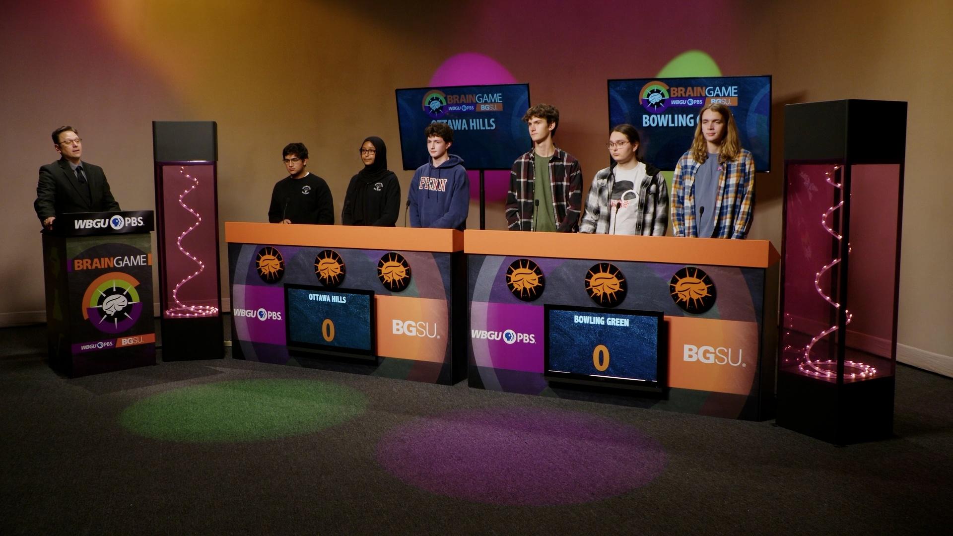 An image from the BGSU Brain Game set. Host Marc Hathaway is behind the podium on the left side and in the center there are two larger podiums- one for Ottawa Hills and one for Bowling Green. There are three students behind each of the larger podiums. Green and purple lights adorn the set.