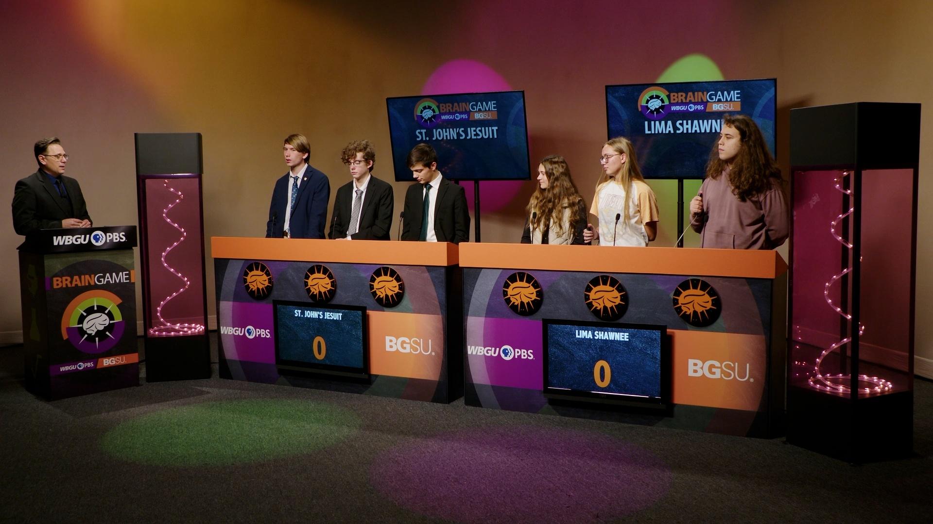 An image from the BGSU Brain Game set. Host Marc Hathaway is behind the podium on the left side and in the center there are two larger podiums- one for St. John’s Jesuit and one for Lima Shawnee. There are three students behind each of the larger podiums. Green and purple lights adorn the set.