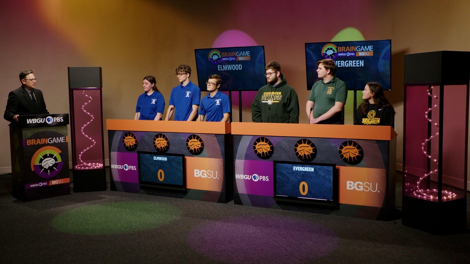 An image from the BGSU Brain Game set. Host Marc Hathaway is behind the podium on the left side and in the center there are two larger podiums- one for Elmwood and one for Evergeen. There are three students behind each of the larger podiums. Green and purple lights adorn the set.