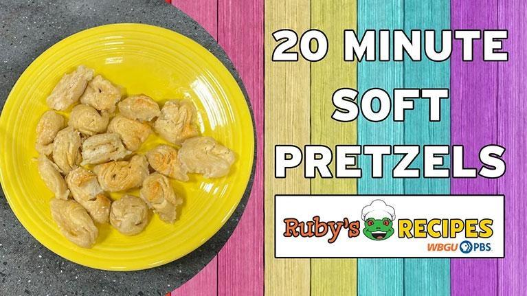 A promotional graphic for "Ruby's Recipes" on WBGU PBS featuring "20 Minute Soft Pretzels." On the left, a bright yellow plate is filled with golden-brown, bite-sized soft pretzel pieces. The right side of the image features a colorful background of vertical wooden planks in pink, yellow, teal, and purple with the recipe title in bold white lettering. At the bottom right is the show's logo, which includes a cartoon frog wearing a chef's hat.