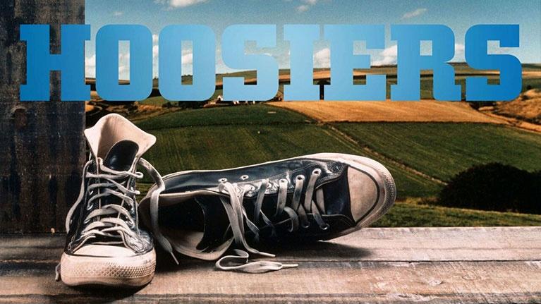 A pair of worn black and white high-top sneakers sits on a wooden surface with a rural landscape and the word "HOOSIERS" in the background.