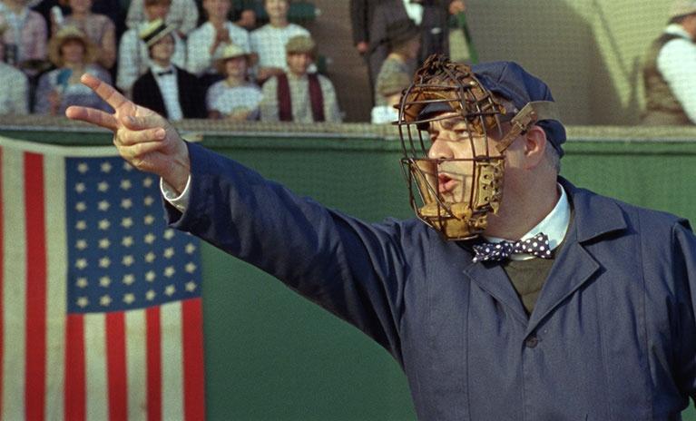 A man dressed as a vintage baseball umpire, wearing a wire cage mask, makes a hand signal during a re-enactment scene from the documentary Signs of the Time. 