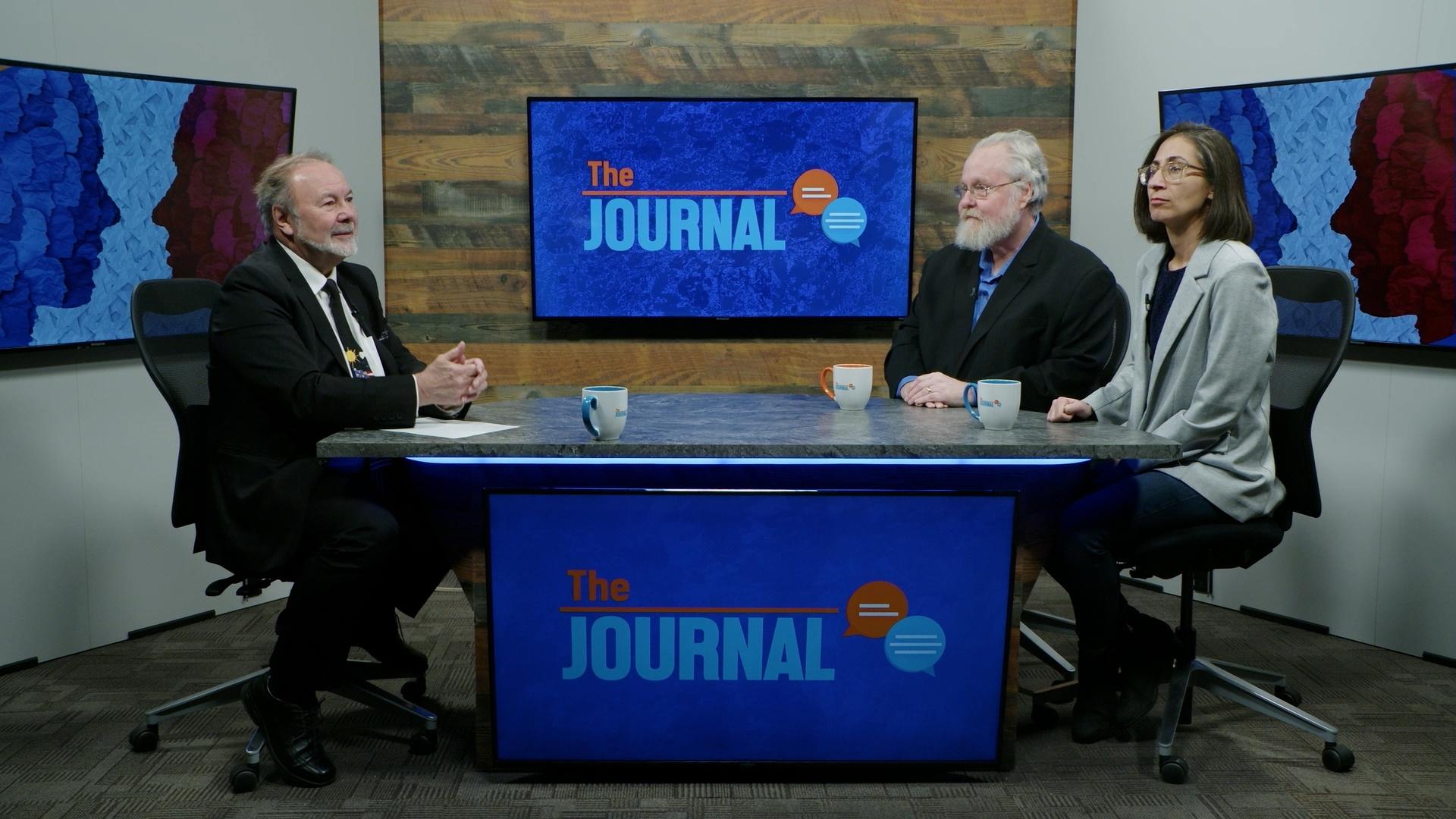 An image from the Journal show set, with host Steve Kendall sitting on the left side and his two guests sitting on the right side. The three are all sitting at a table in front of monitors that have the show logo.