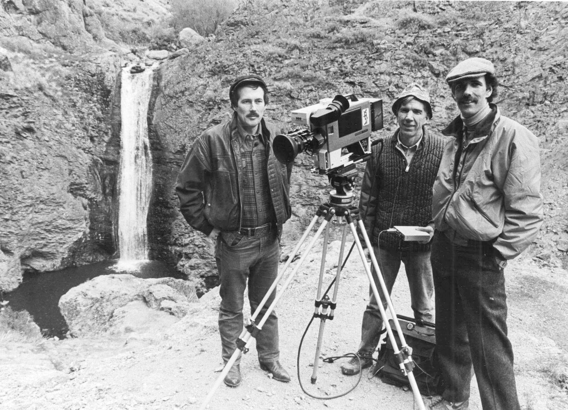 Three men standing next to a small waterfall with a video camera