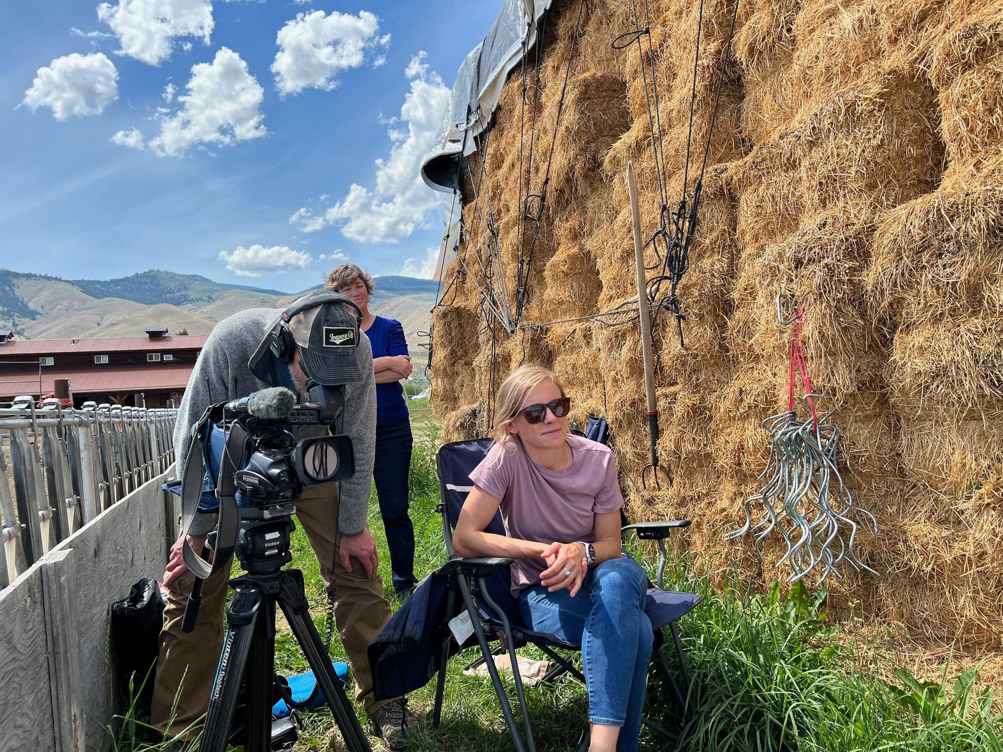 A woman sitting next to a large hay bale while someone films next to her.