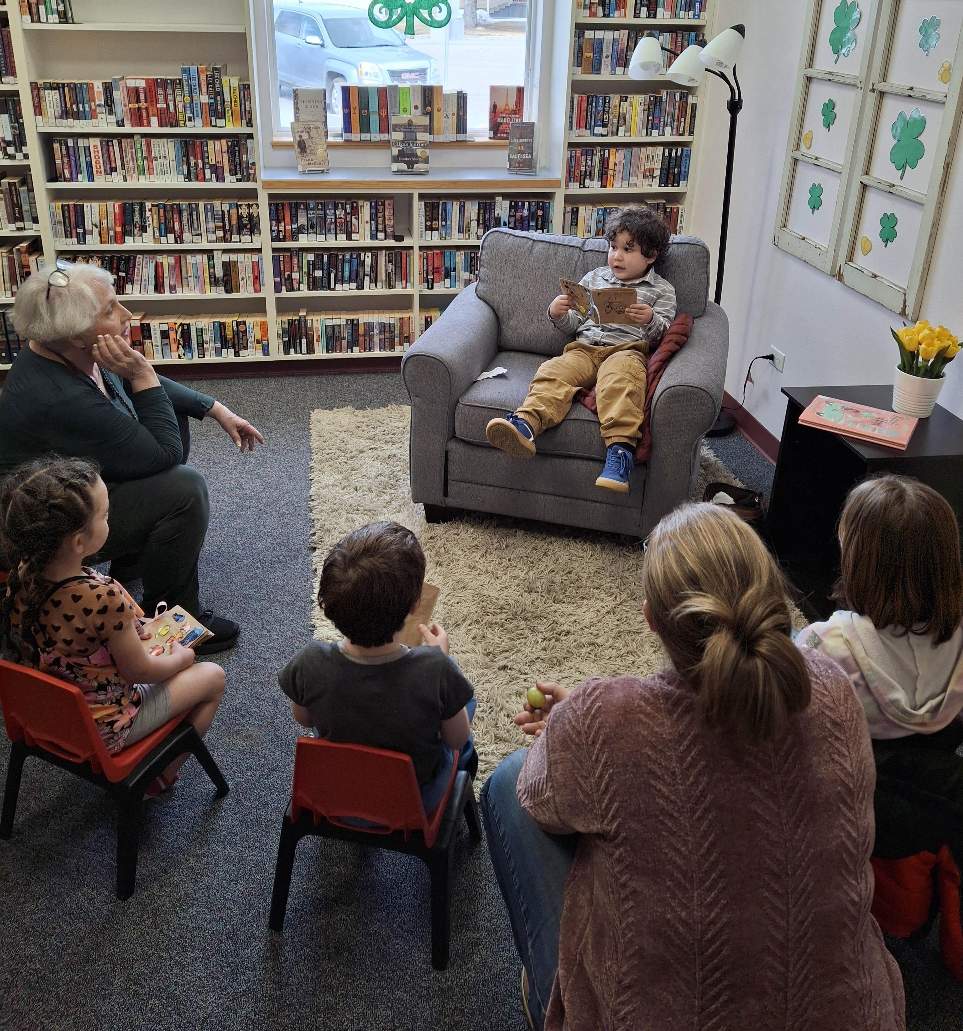 A kid sits on a seat and reads his book to a group