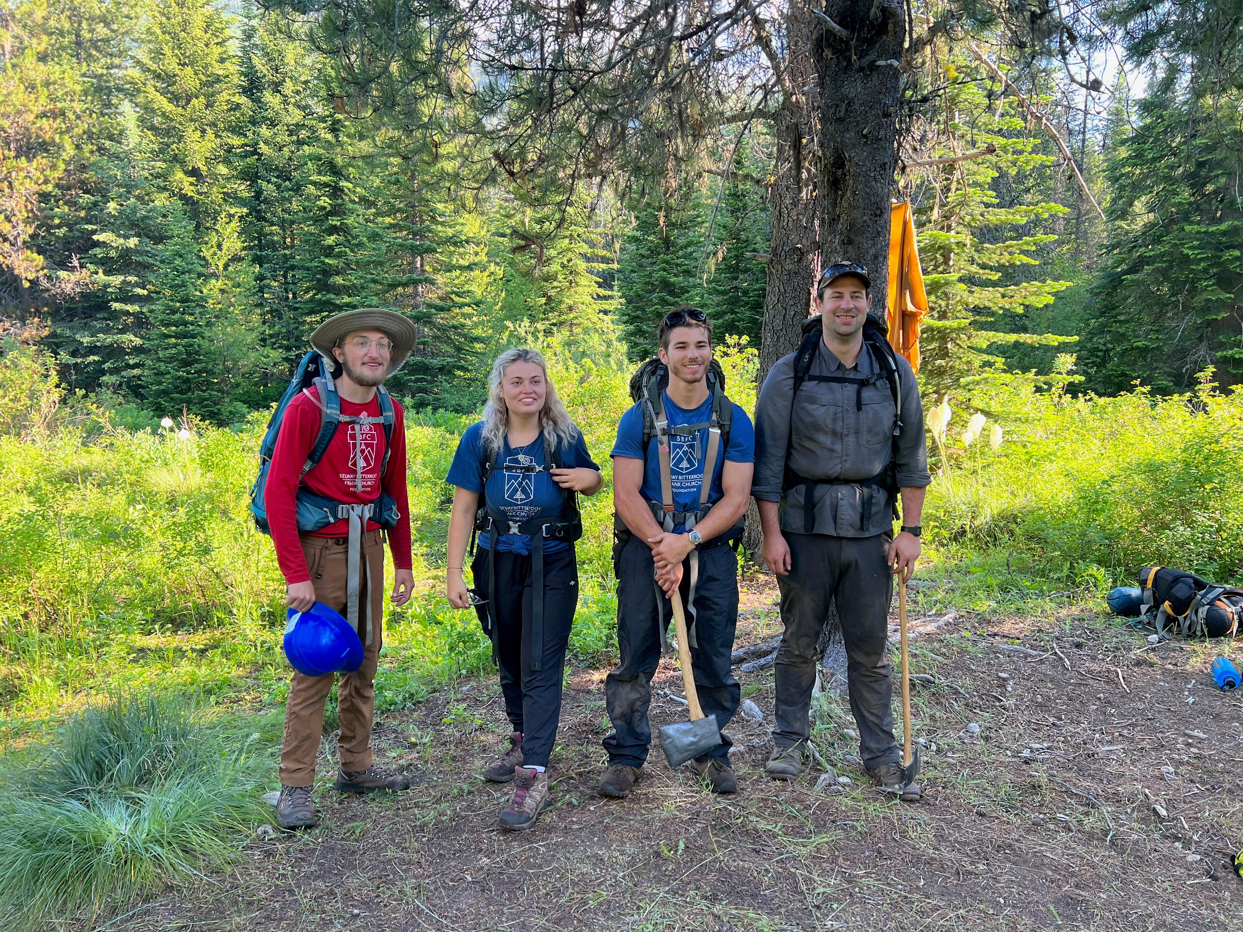 Four hikers stand together and smile. Two of them are holding axes