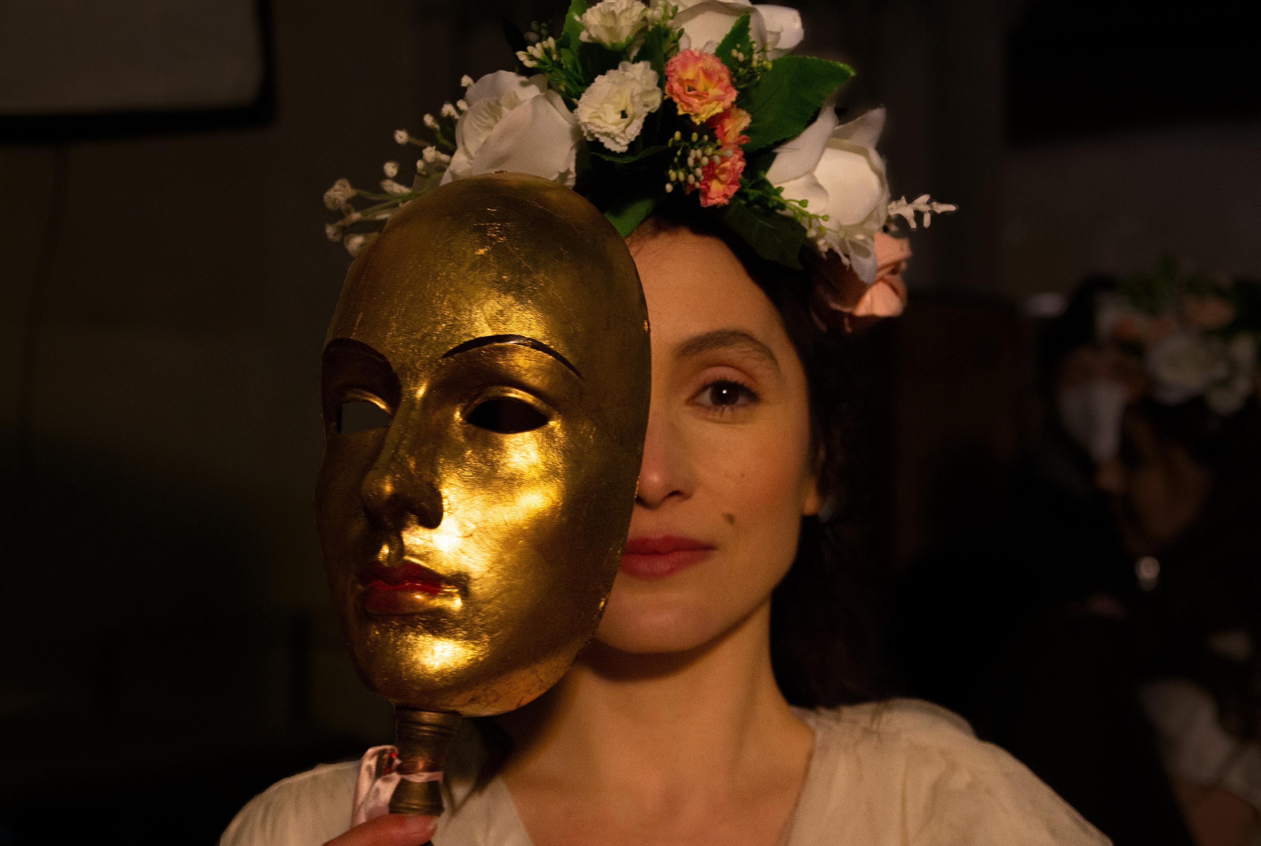 Photo of a woman showing half of her face behind a gold mask with an ornate flower arrangement in her hair.