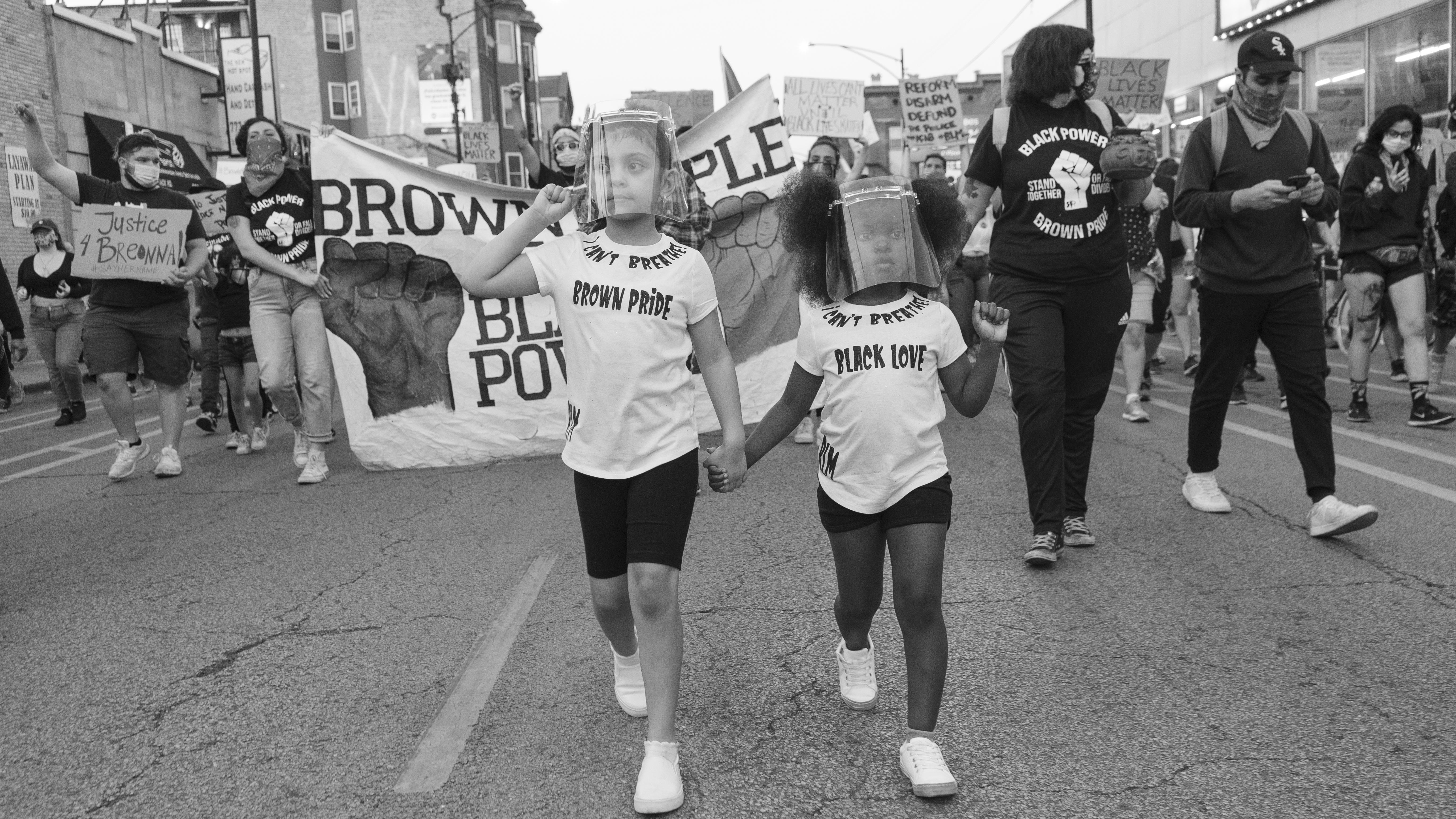 Protestors gather on a street with two young girls at the forefront, wearing shirts that say "brown pride" and "Black love"