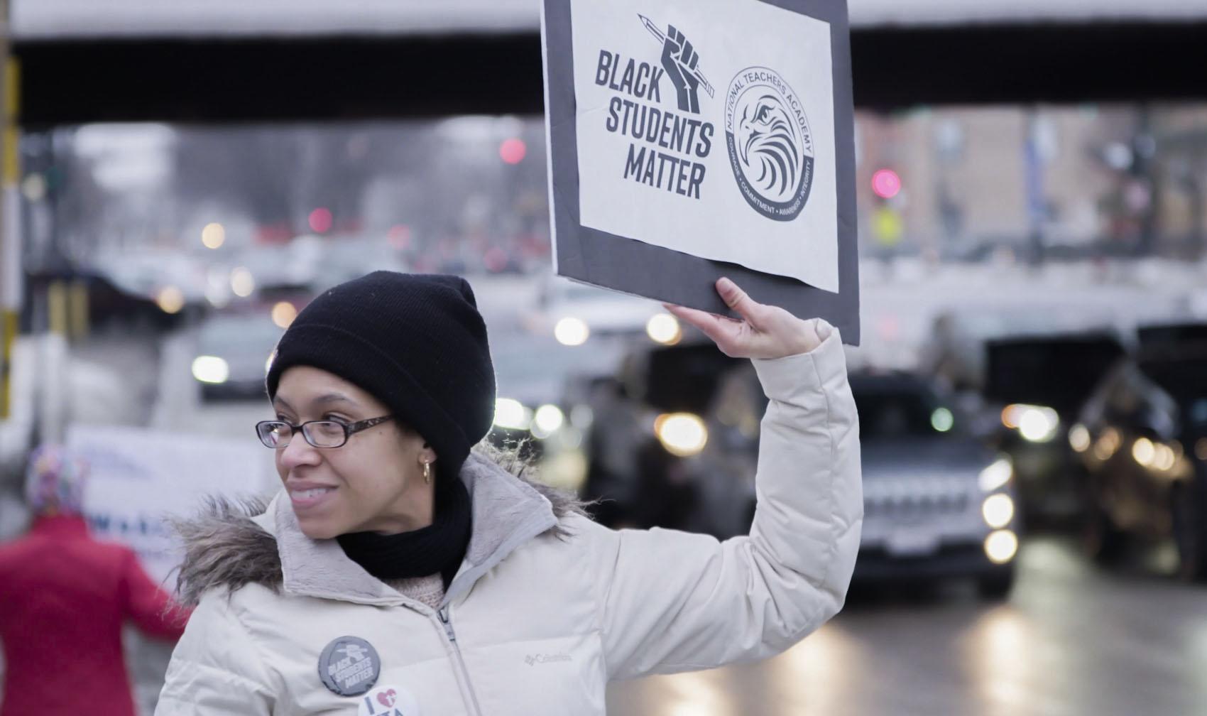 A woman in winter clothes hold up a "Black Students Matter" sign on the side of a busy Chicago street.