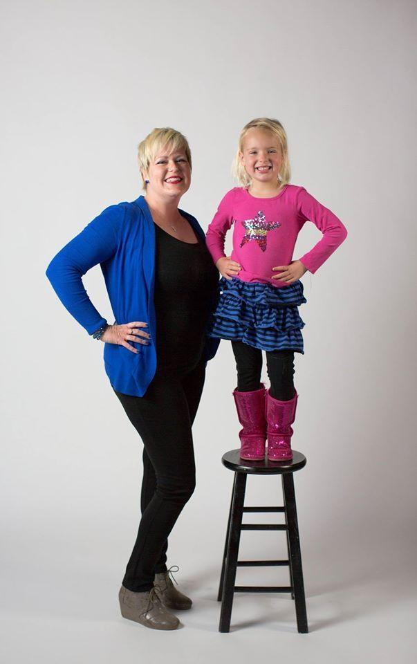 Photo of a woman smiling as her young daughter stands next to her on a stool. 