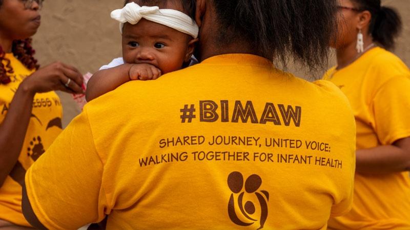 Woman holding infant, woman wears yellow #BIMAW T-Shirt.