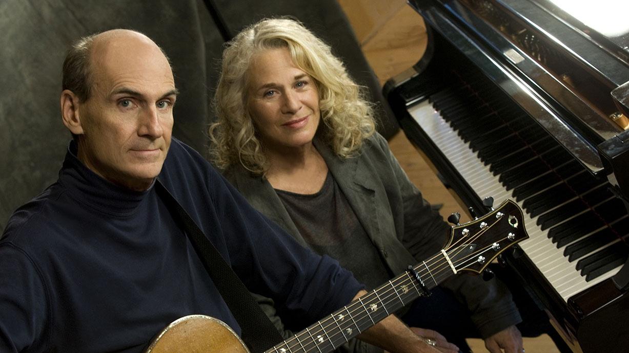 James Taylor holds a guitar and sits next to Carole King, who is at a Piano
