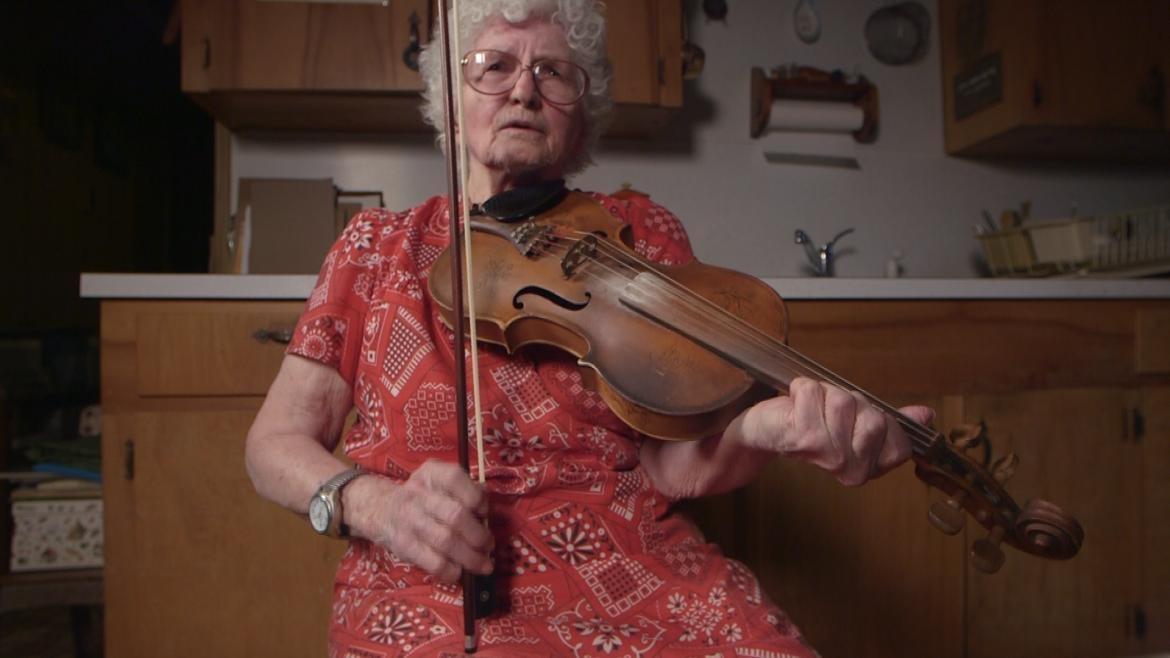 A woman plays her fiddle in her kitchen.