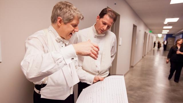 Photo of two men talking in a hallway while holding sheet music