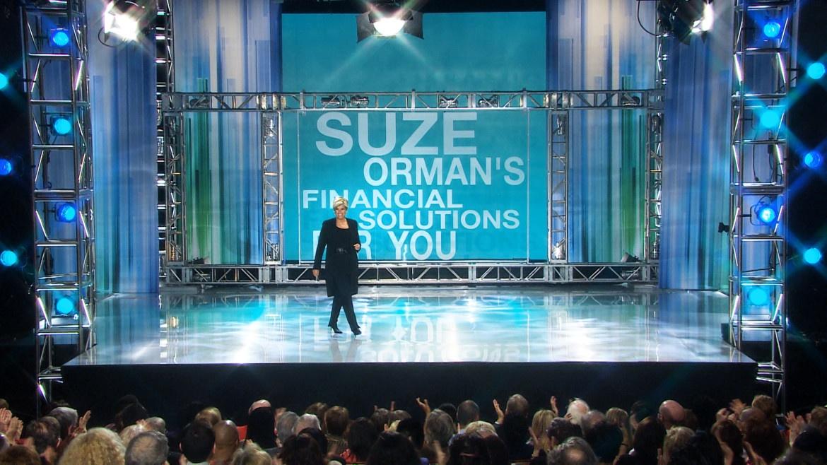 PHoto of a woman on stage with "Suze Orman's Financial Solutions for You" sign in background