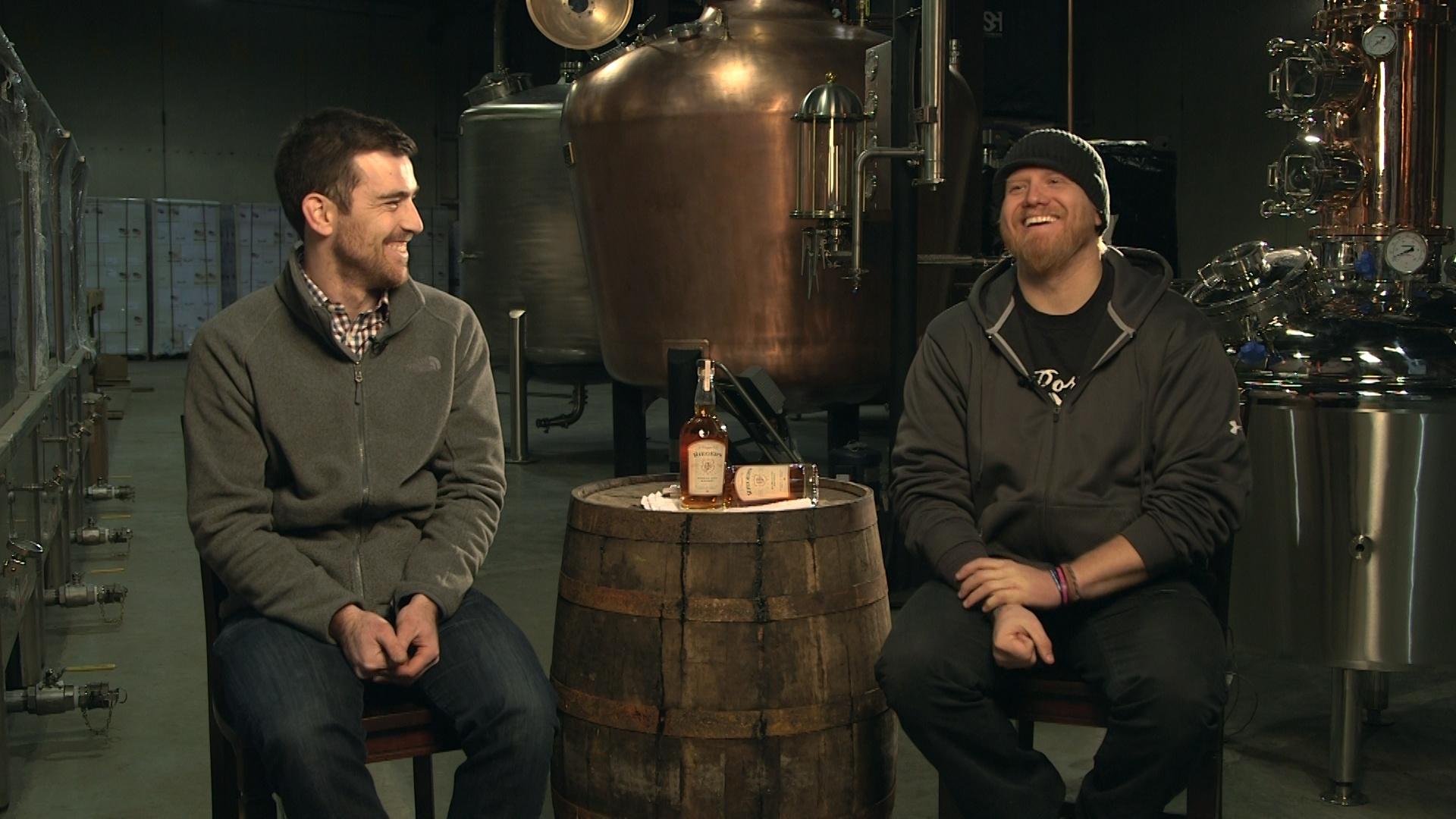 Two men sit in a distillery laughint. A barrel sits between them, and two bottles of alcohol are on the barrel. 