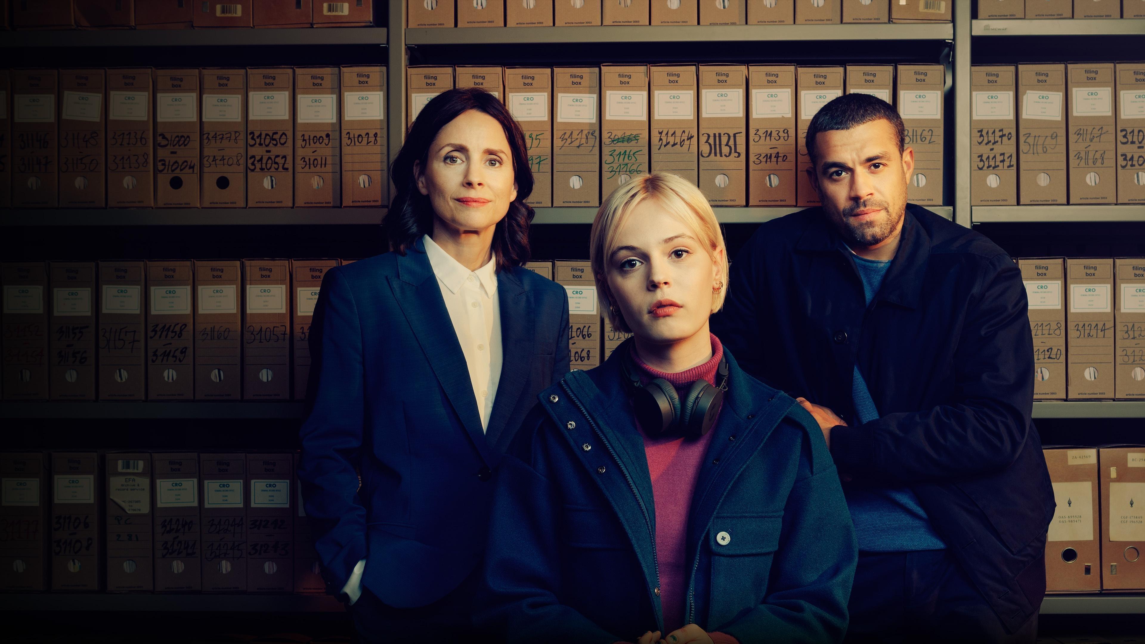 The main character from the PBS television show Patience is shown, wearing headphones and a colorful sweater, sitting at a table. They are holding a small wooden dinosaur skeleton model, which is highlighted by a red arrow. The background includes a lamp and part of the room's wall.