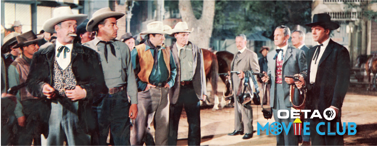A dramatic scene from Gunfight at the O.K. Corral (1957) featuring a tense standoff between two groups of men in Old West attire outside the “Ranch Saloon.” On the right, suited lawmen and gunfighters—including characters representing Wyatt Earp and Doc Holliday—stand armed and ready. On the left, townsmen and cowboys in hats and vests watch anxiously. The image captures the iconic tension leading up to the legendary shootout in Tombstone, Arizona.