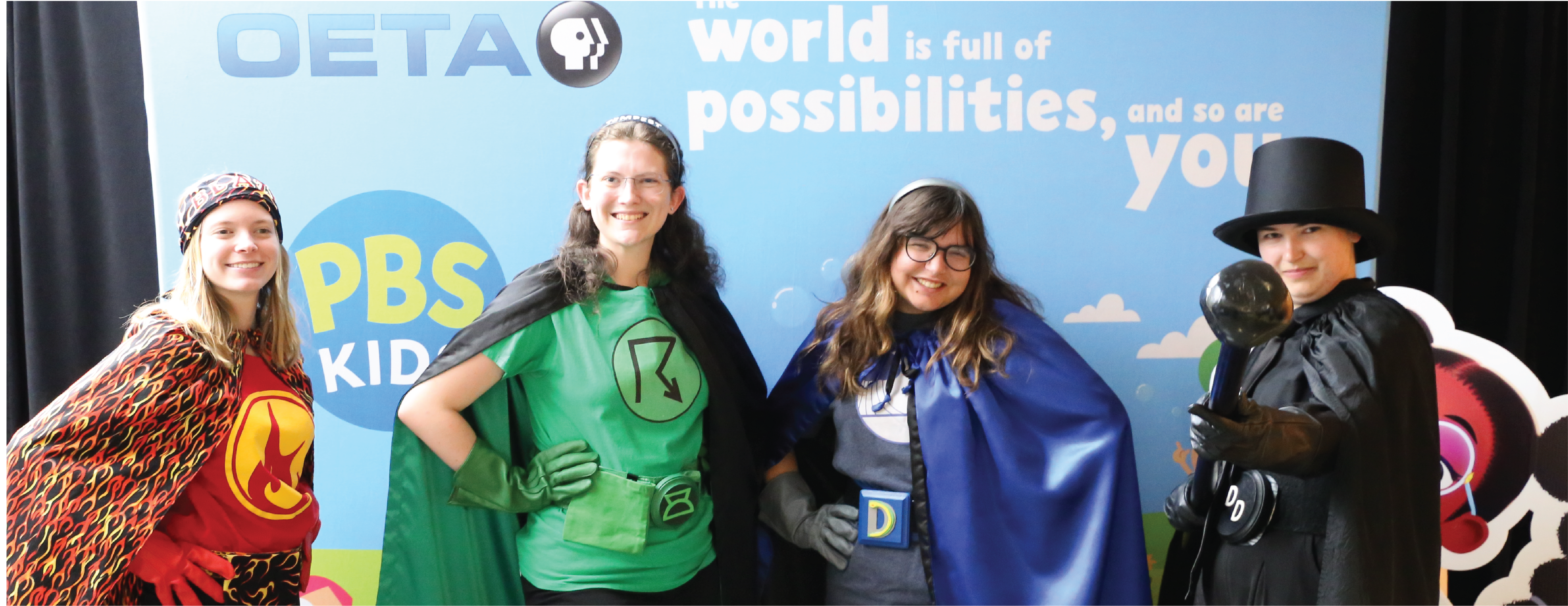 Four smiling adults in superhero costumes representing different weather types pose in front of a PBS Kids and OETA backdrop that reads ‘The world is full of possibilities, and so are you.
