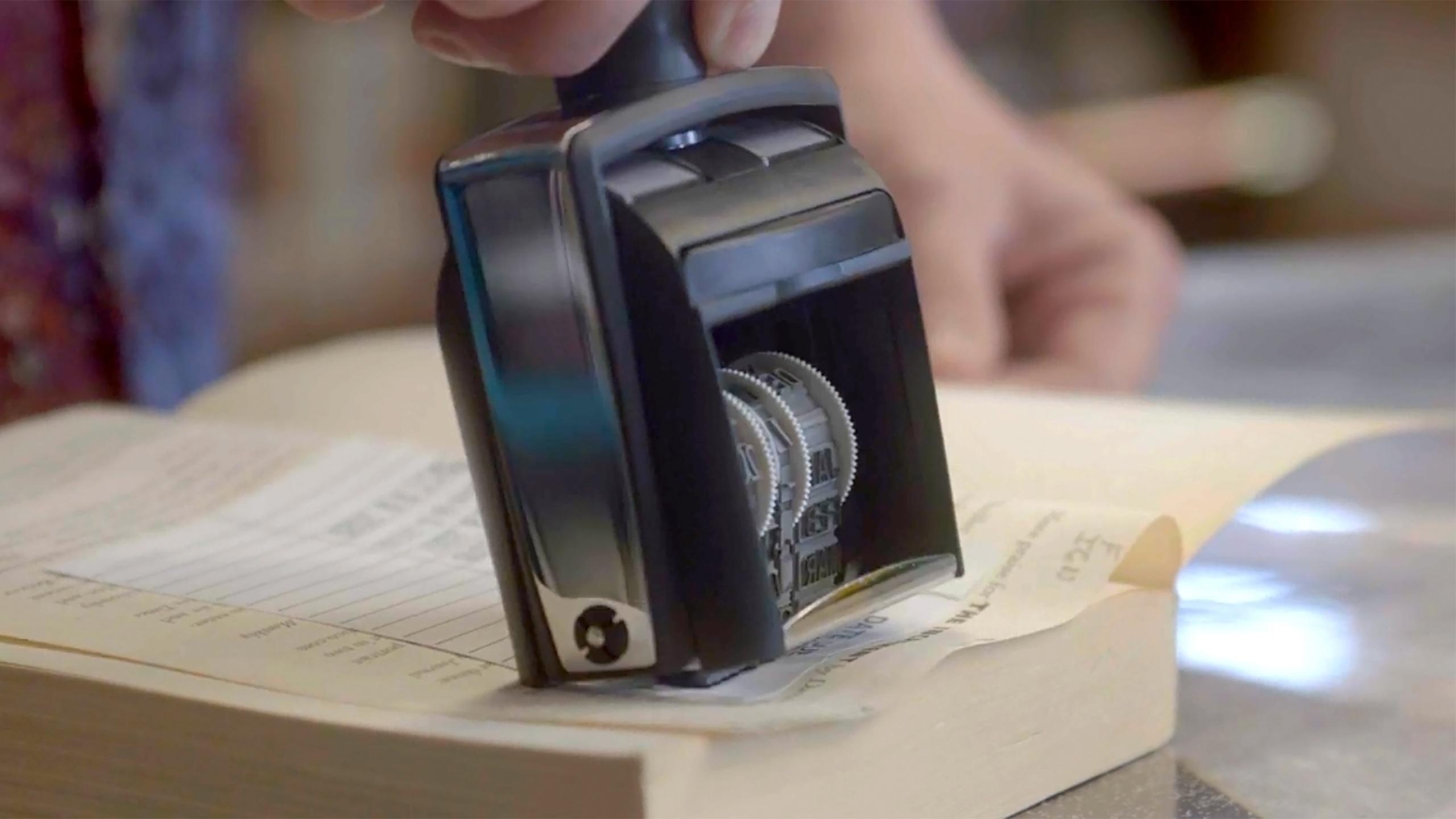 A black and silver “due by” stamper marks a date inside an open book, with a hand holding the book open in the background. 