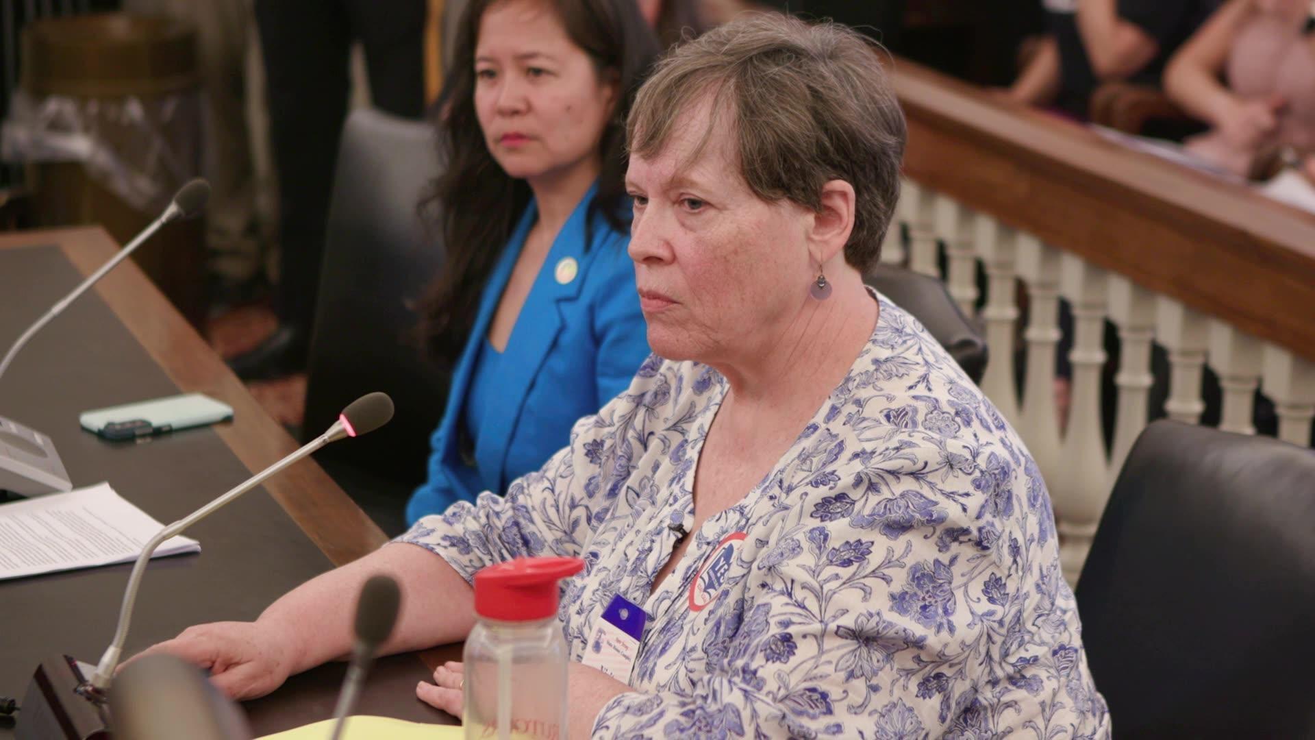 A woman with short brown hair sits at a courtroom table in front of a microphone, serious, with another woman beside her.