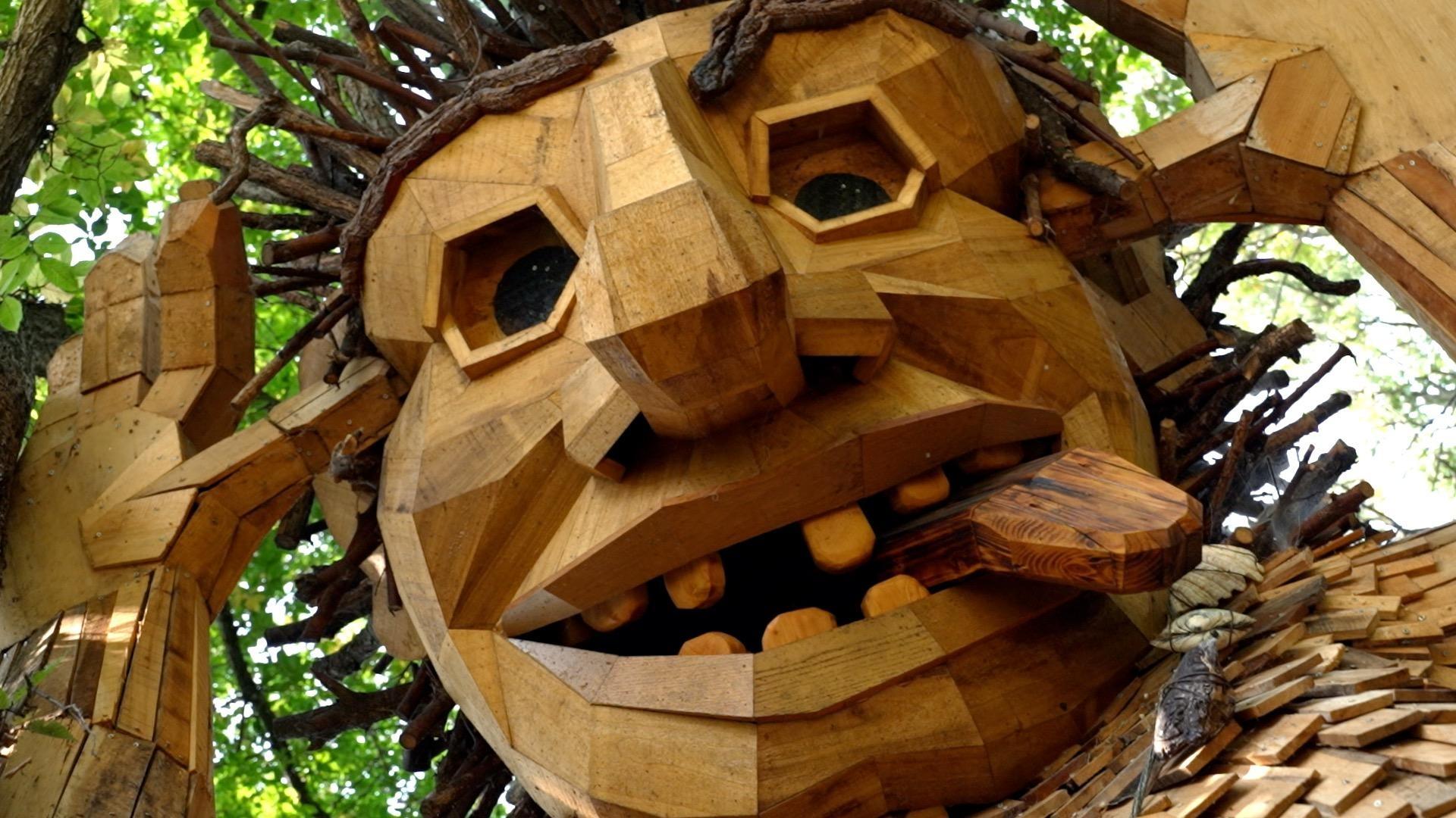 A close-up of a large outdoor wooden troll sculpture with a geometric, faceted face, hexagonal eyes, and an open mouth with rounded wooden teeth. The figure's hair is made from branches and twigs, and it is nestled among green trees.