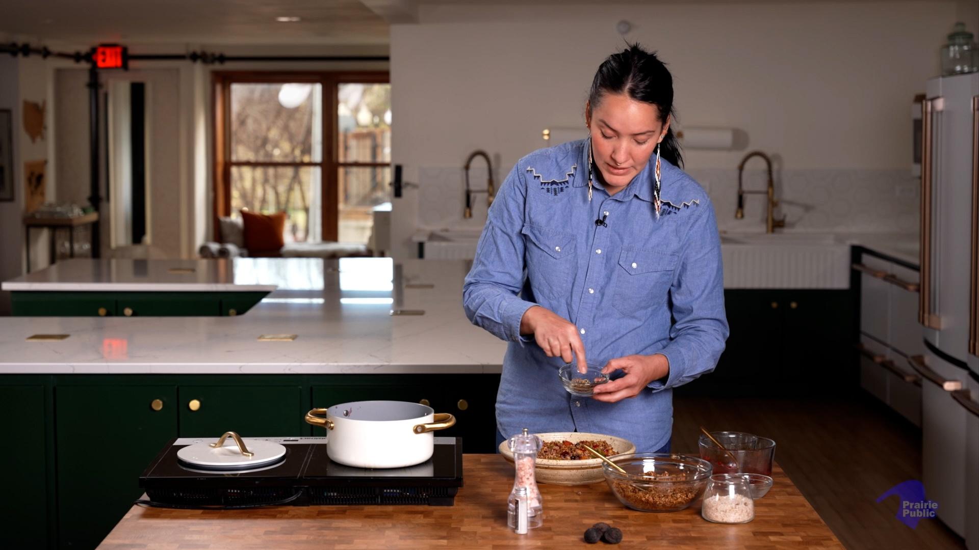 A woman wearing a blue denim shirt and beaded earrings adds seasoning to a bowl of food at a butcher block counter in a professional kitchen. A white Dutch oven sits on an induction cooktop nearby, alongside bowls of ingredients.