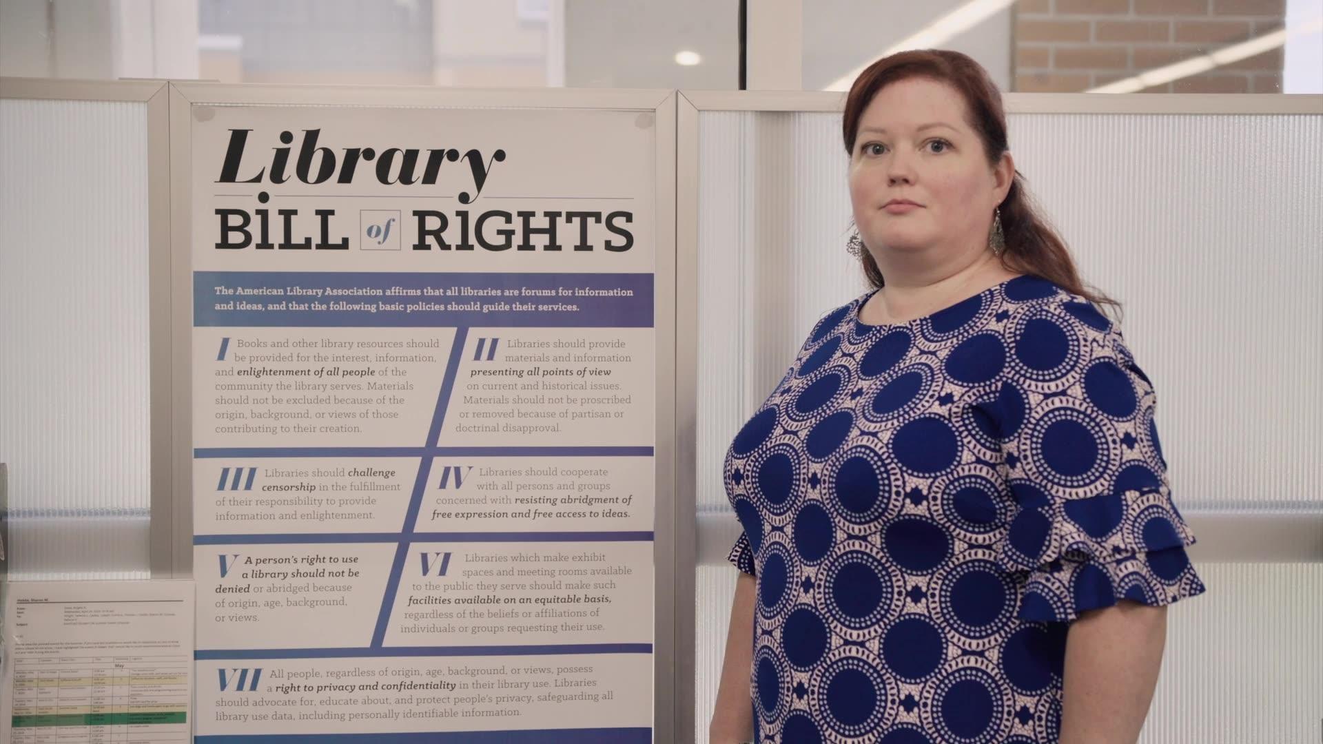 A woman with red hair in a blue and white patterned shirt stands next to a Library Bill of Rights poster with black text and blue Roman numerals on a white background
