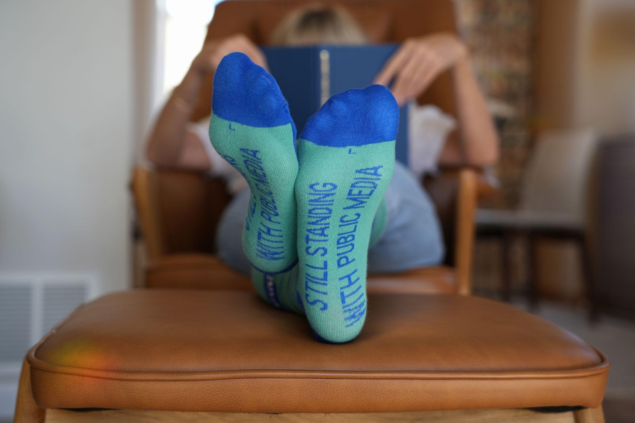 A woman wears a pair of socks that say "Still Standing With Public Media" on the sole.