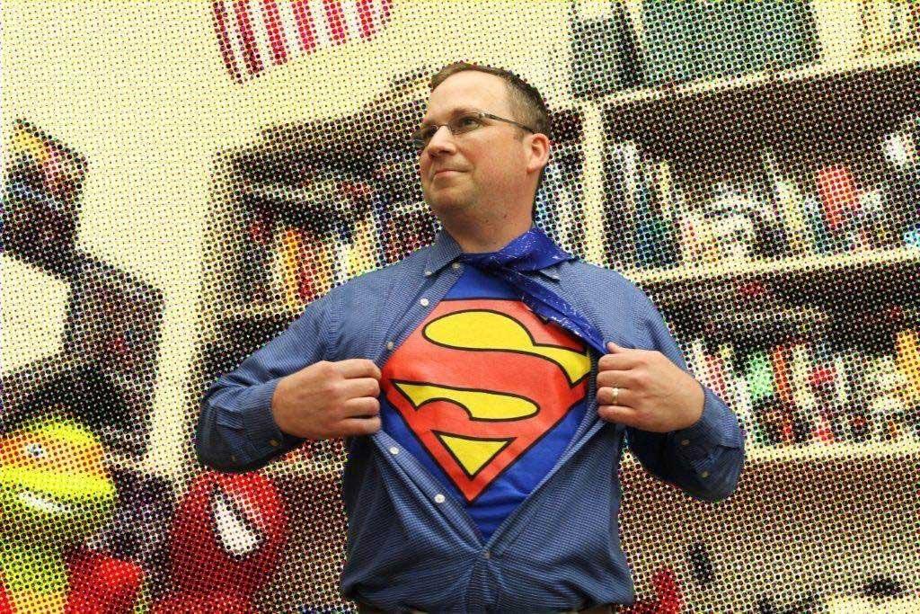 A teacher with glasses opens his shirt to reveal a Superman logo underneath